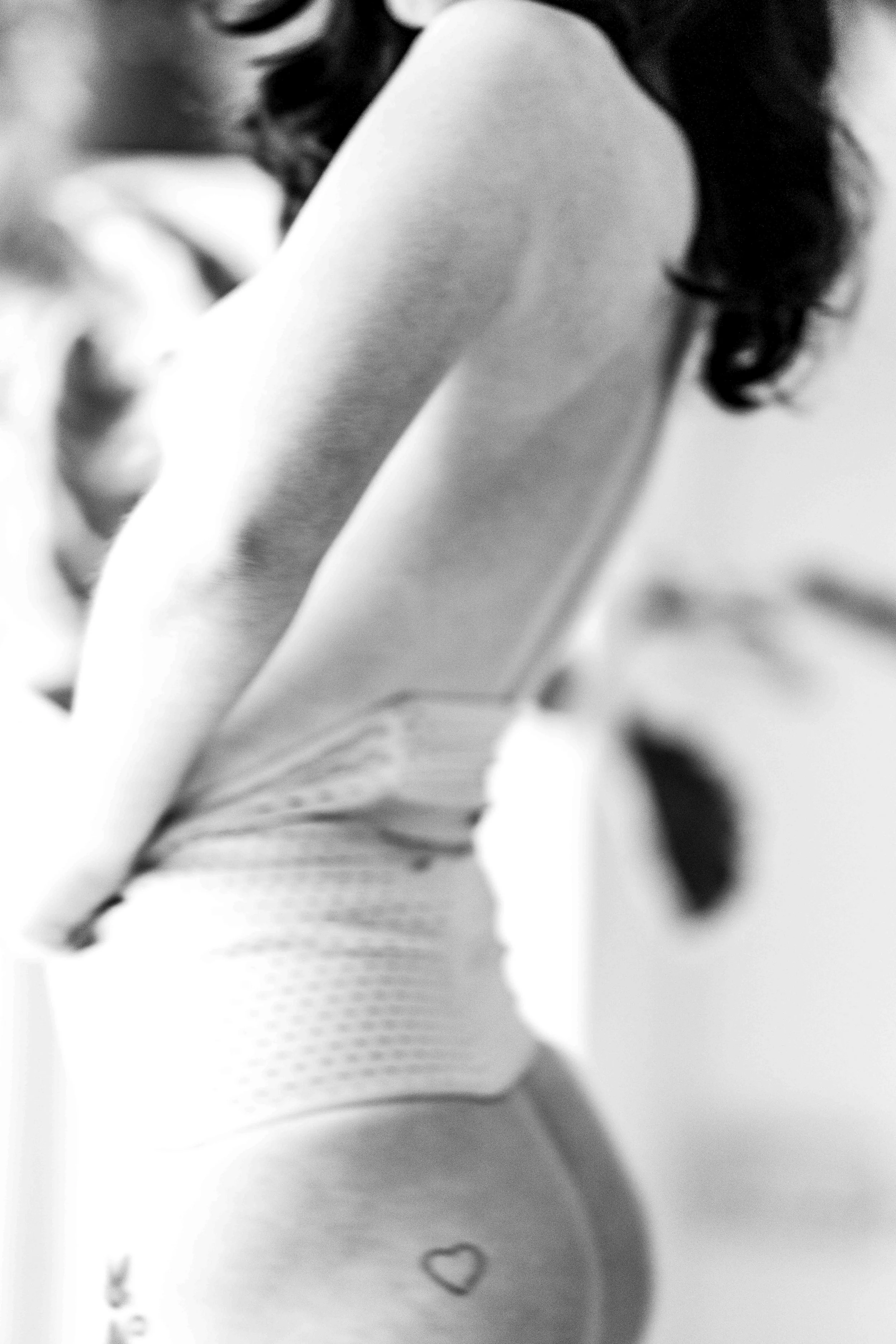 Close-up black and white photo of a woman's torso, with her wearing a sleeveless top and fishnet leggings, revealing a small heart tattoo on her side.