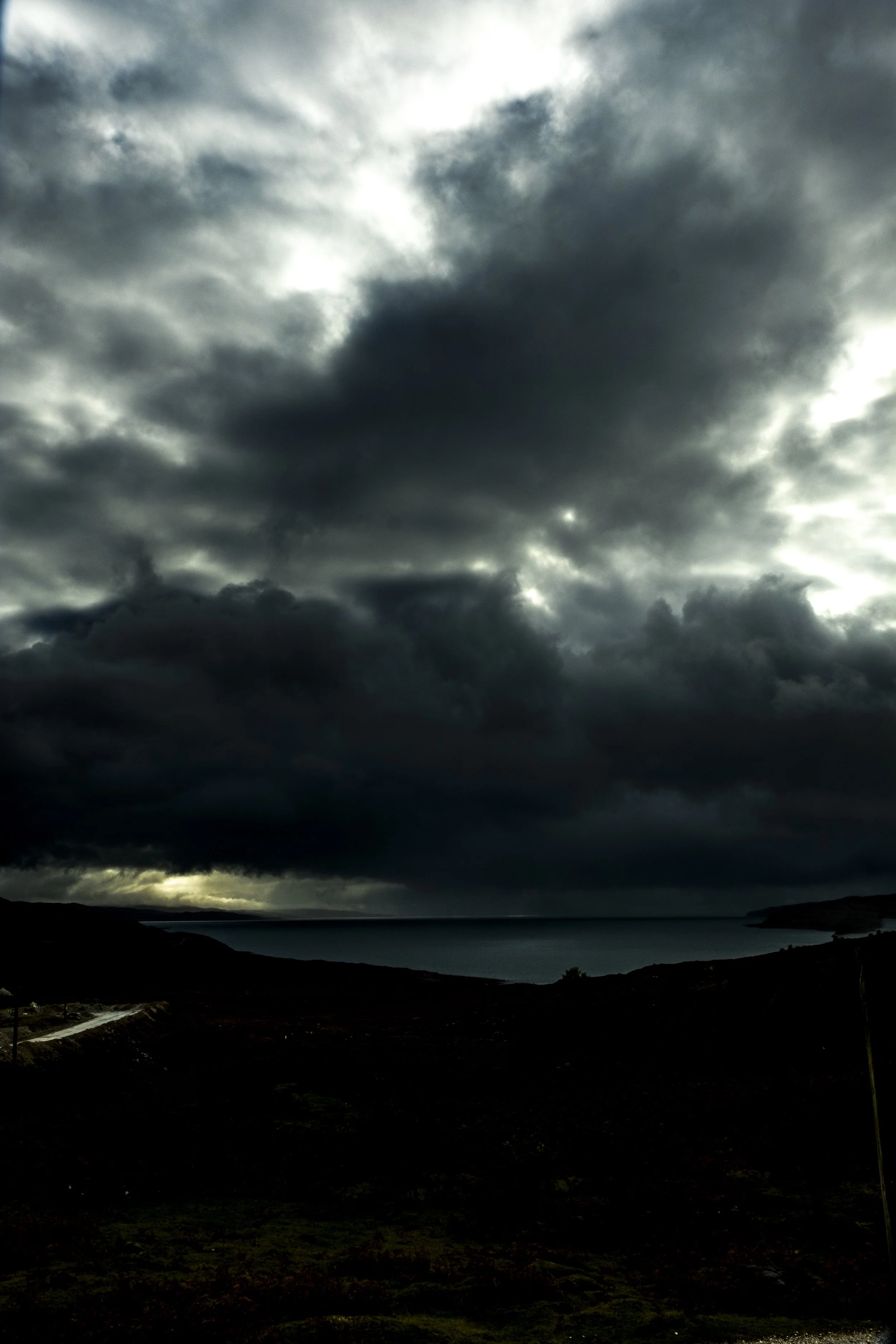 Dark storm clouds gather over a body of water with a small landmass or peninsula in the distance. The scene appears ominous with heavy clouds and limited visibility.
