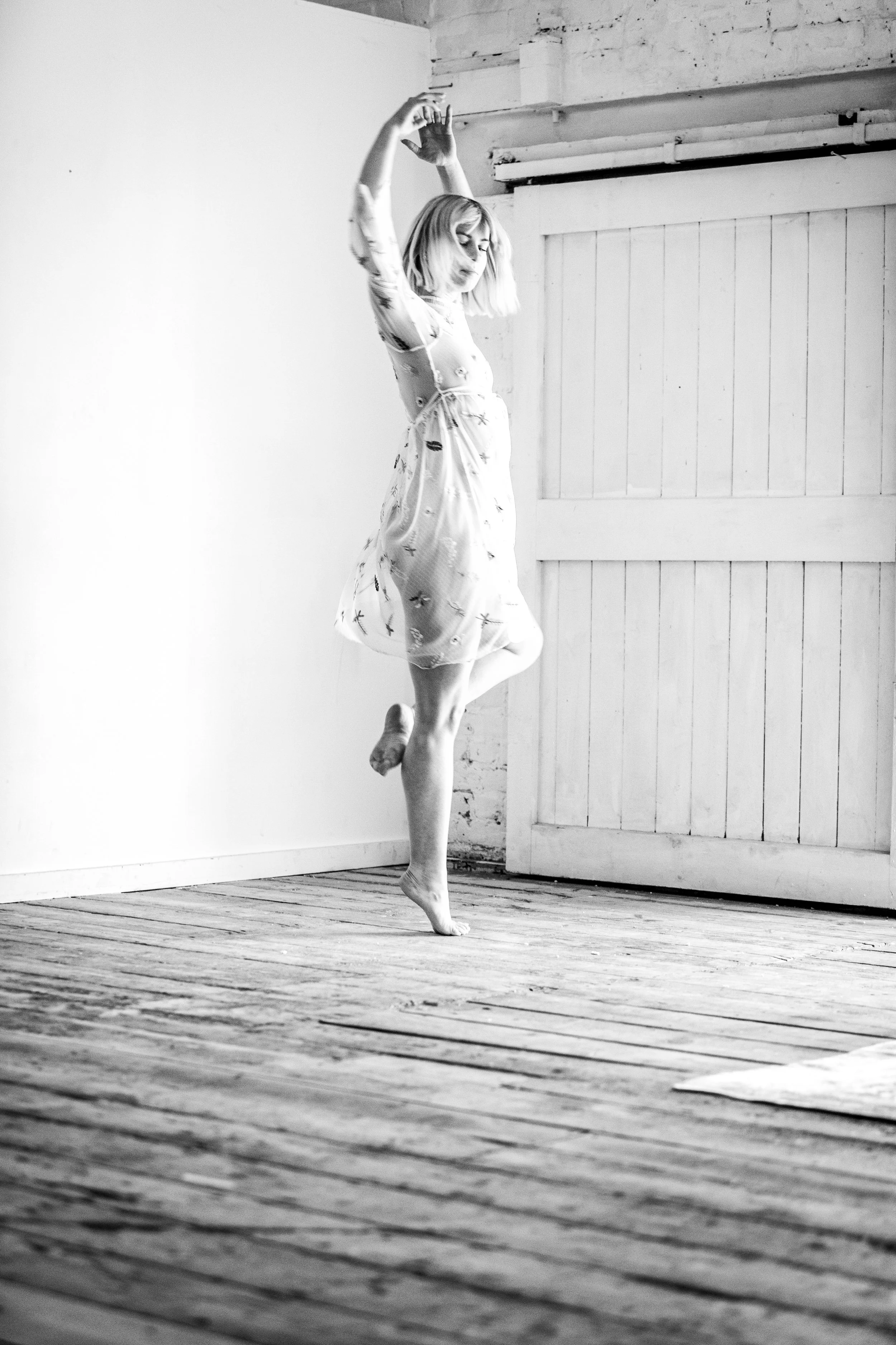 A woman with blonde hair performing a ballet pose in a dance studio with wooden floors and a partially open door.