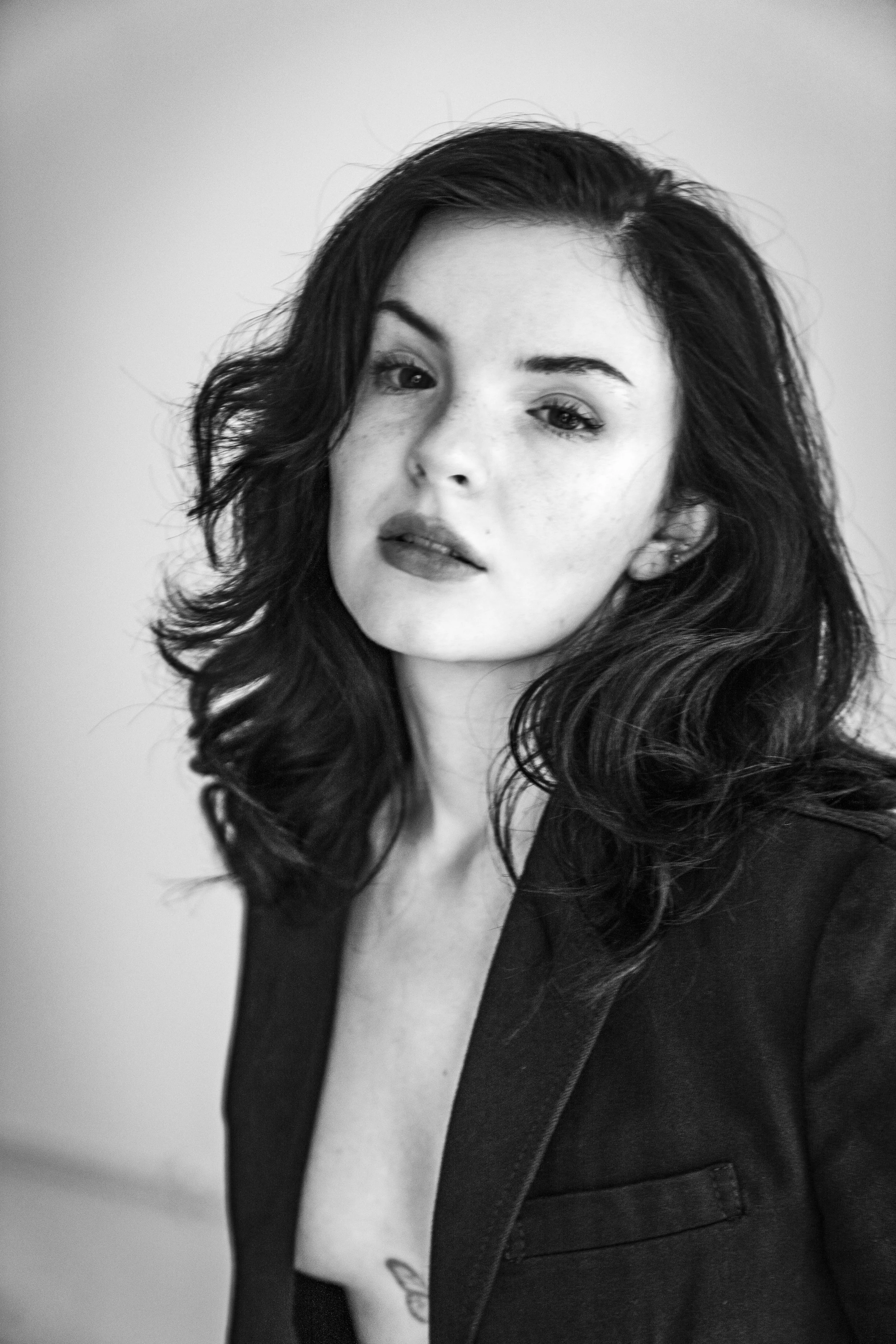 Black and white portrait of a woman with wavy hair wearing a blazer, with a tattoo visible on her chest.