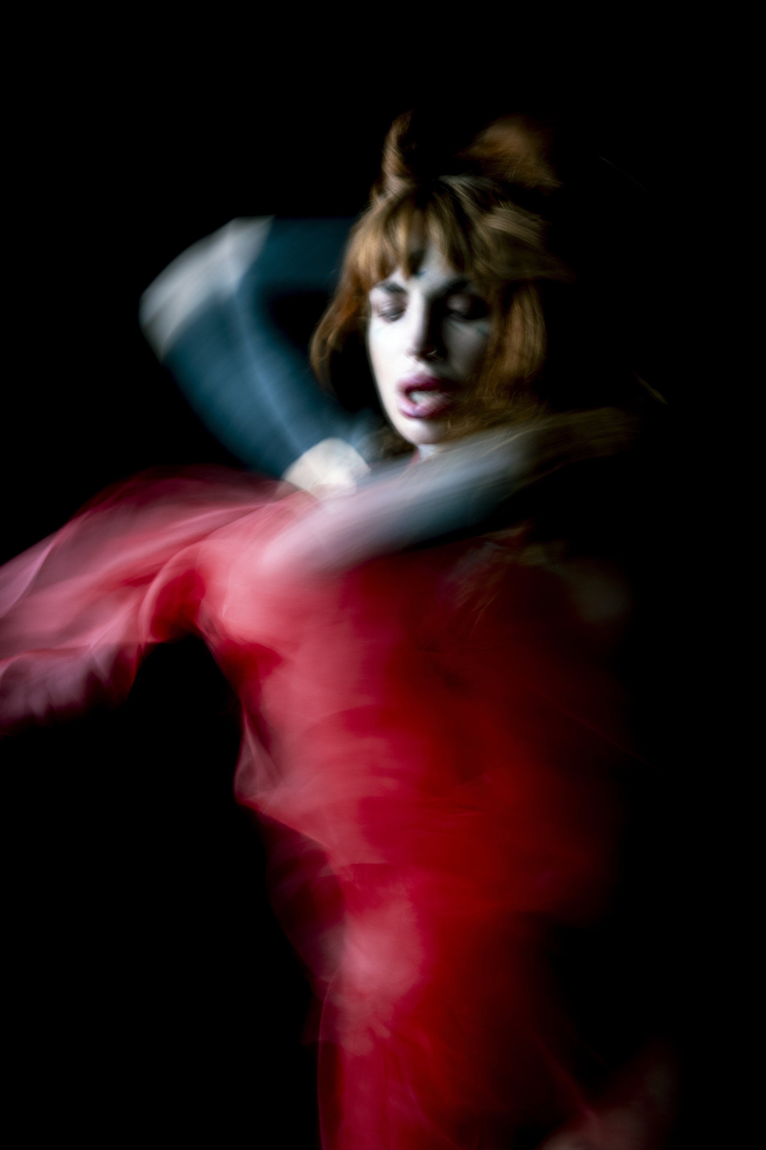 A woman with red hair dancing passionately with blurred motion, wearing a flowing red dress, set against a dark background.