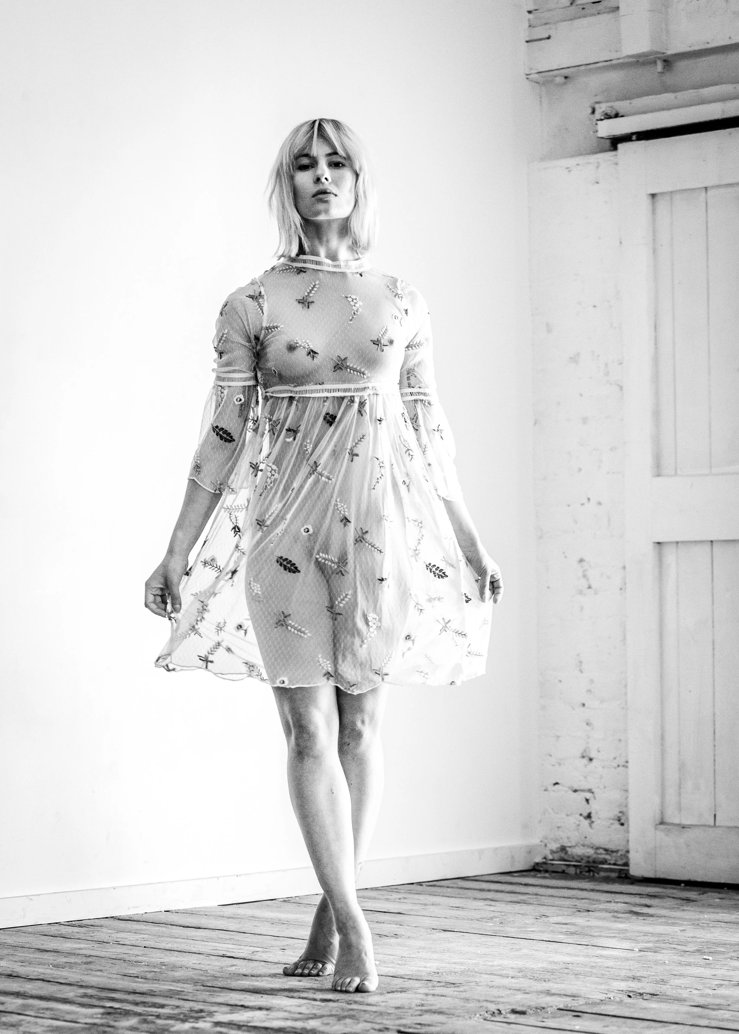 A woman with short blonde hair standing on a wooden floor indoors. She is wearing a sheer dress with embroidered patterns and has a serious expression on her face. The background features a plain wall and a partially visible wooden shelf.