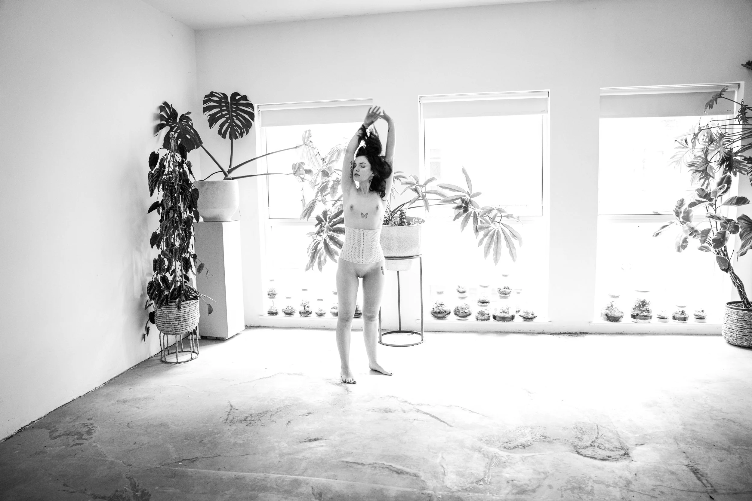 A woman with long dark hair, standing topless with closed eyes, arms raised above her head, in a brightly lit room with large windows, surrounded by potted plants.