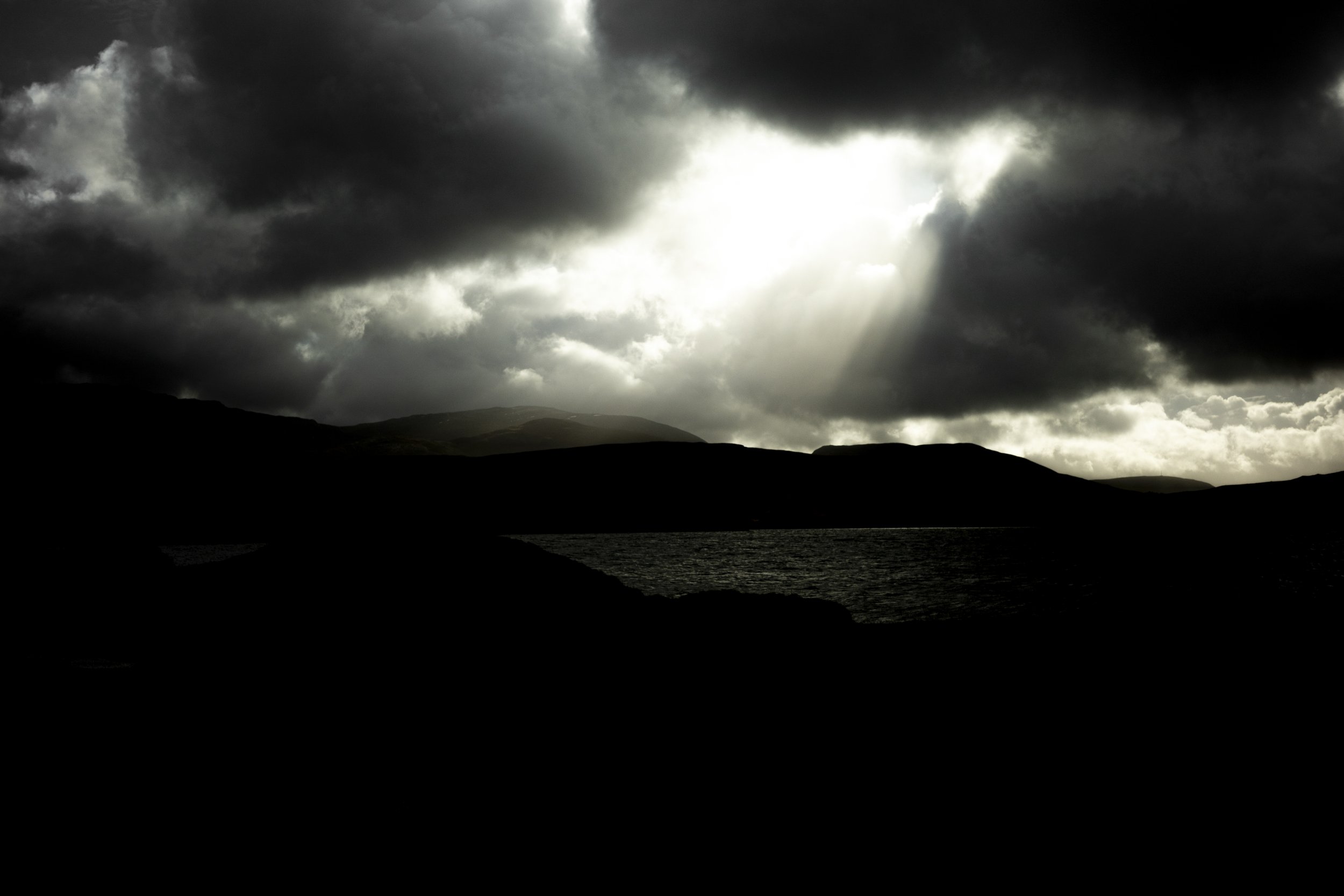 Dark clouds over mountains and a body of water, with sunlight breaking through the clouds.