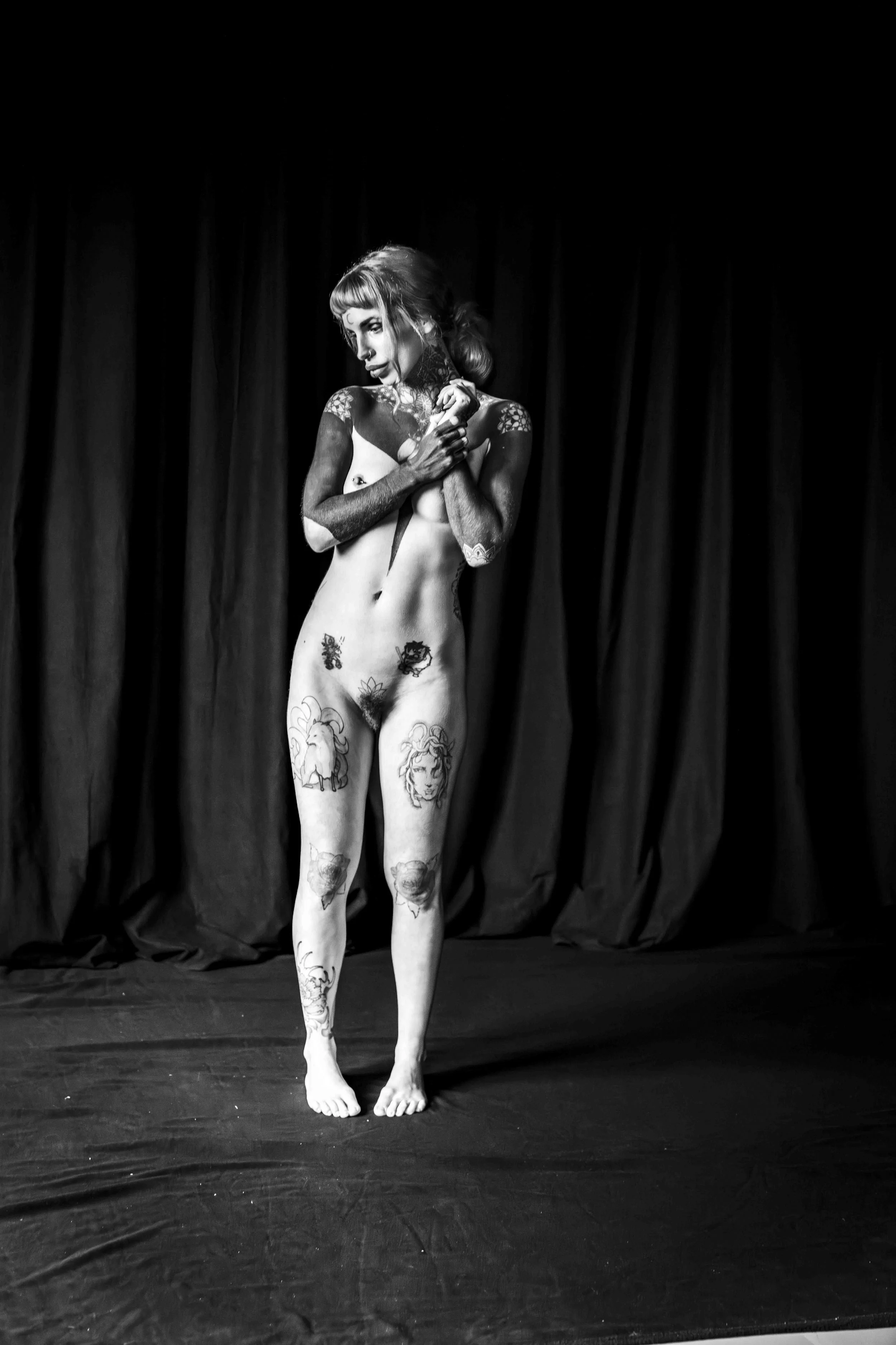 Black and white photo of a woman standing on a stage with a curtain background. She is nude with tattoos on her arms, legs, and torso, and her pose suggests a performance or artistic expression.