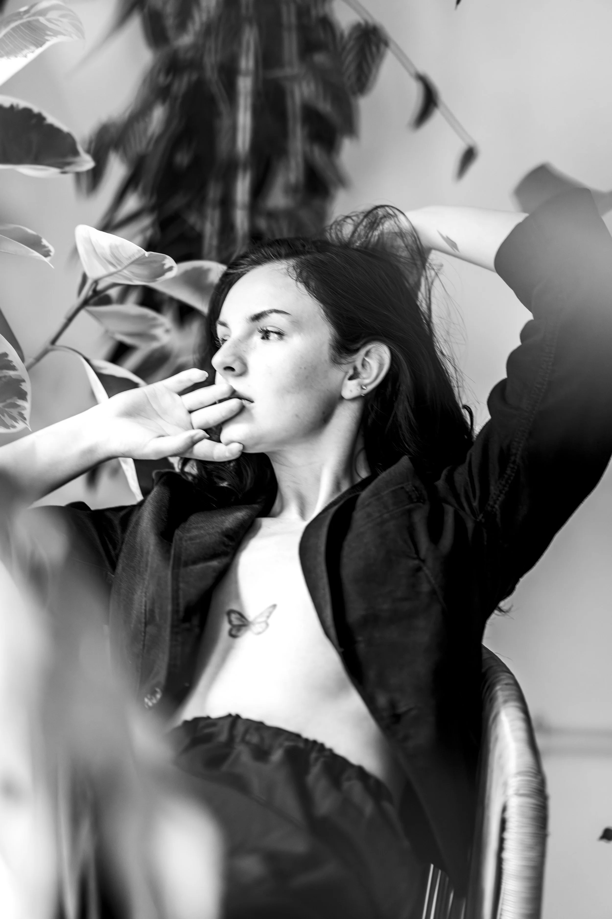 A black and white photo of a young woman with dark hair, looking to the side with a contemplative expression, surrounded by large leafy plants and a chair.