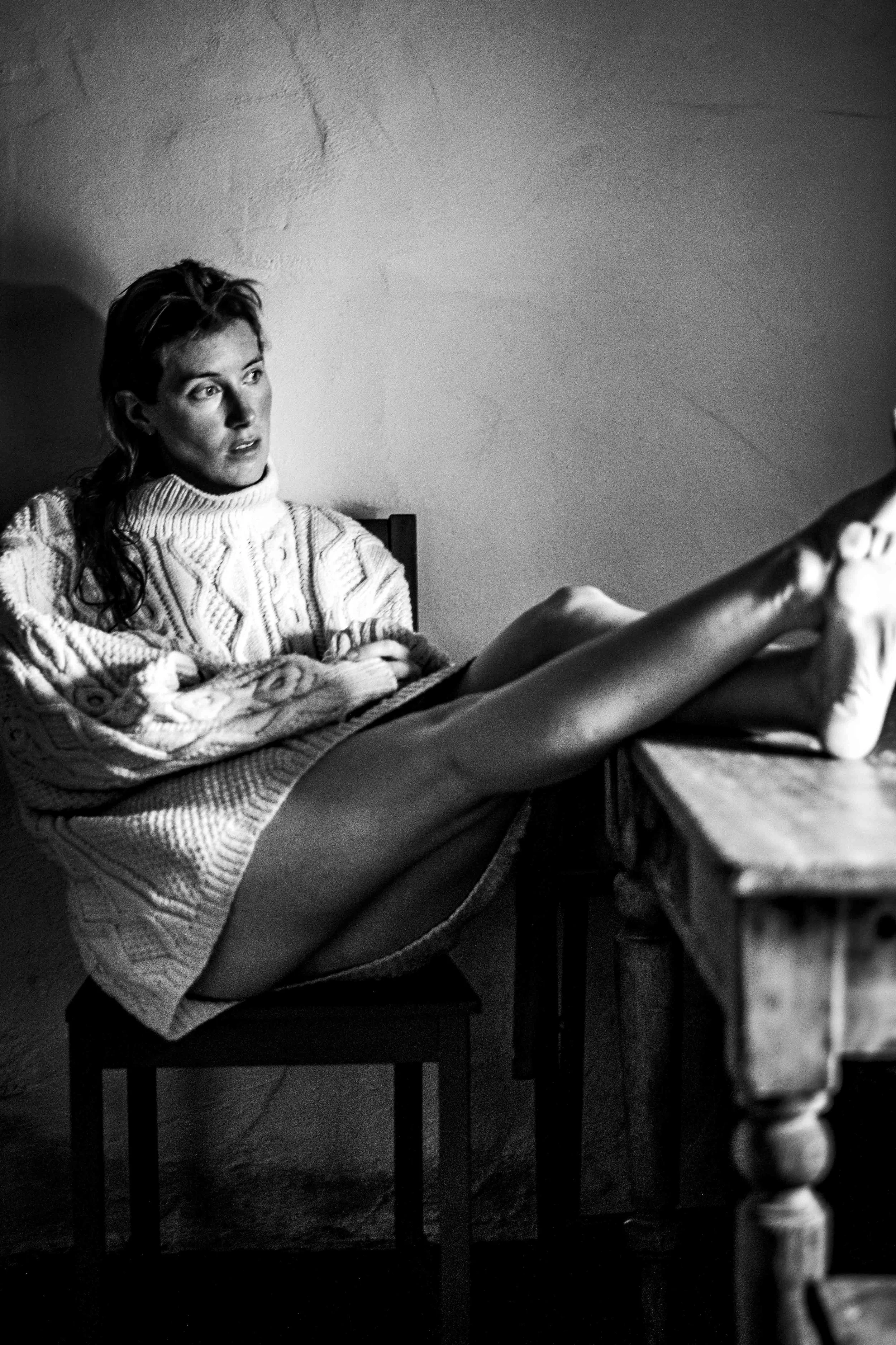 A woman with short hair is sitting on a chair, wearing a cozy, patterned sweater, and looking contemplative. She has her arms crossed on her lap, and her legs are extended on a wooden table. The setting is indoors with a plain, textured wall in the background, captured in black and white.