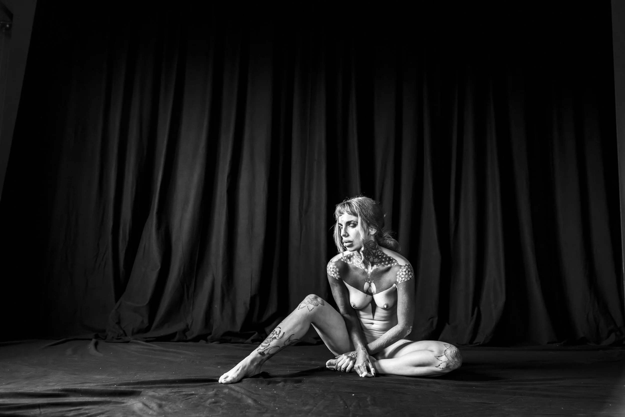 A woman with tattoos and tattoos on her face, wearing minimal clothing, sitting on the floor in front of a black curtain, in a dramatic pose.