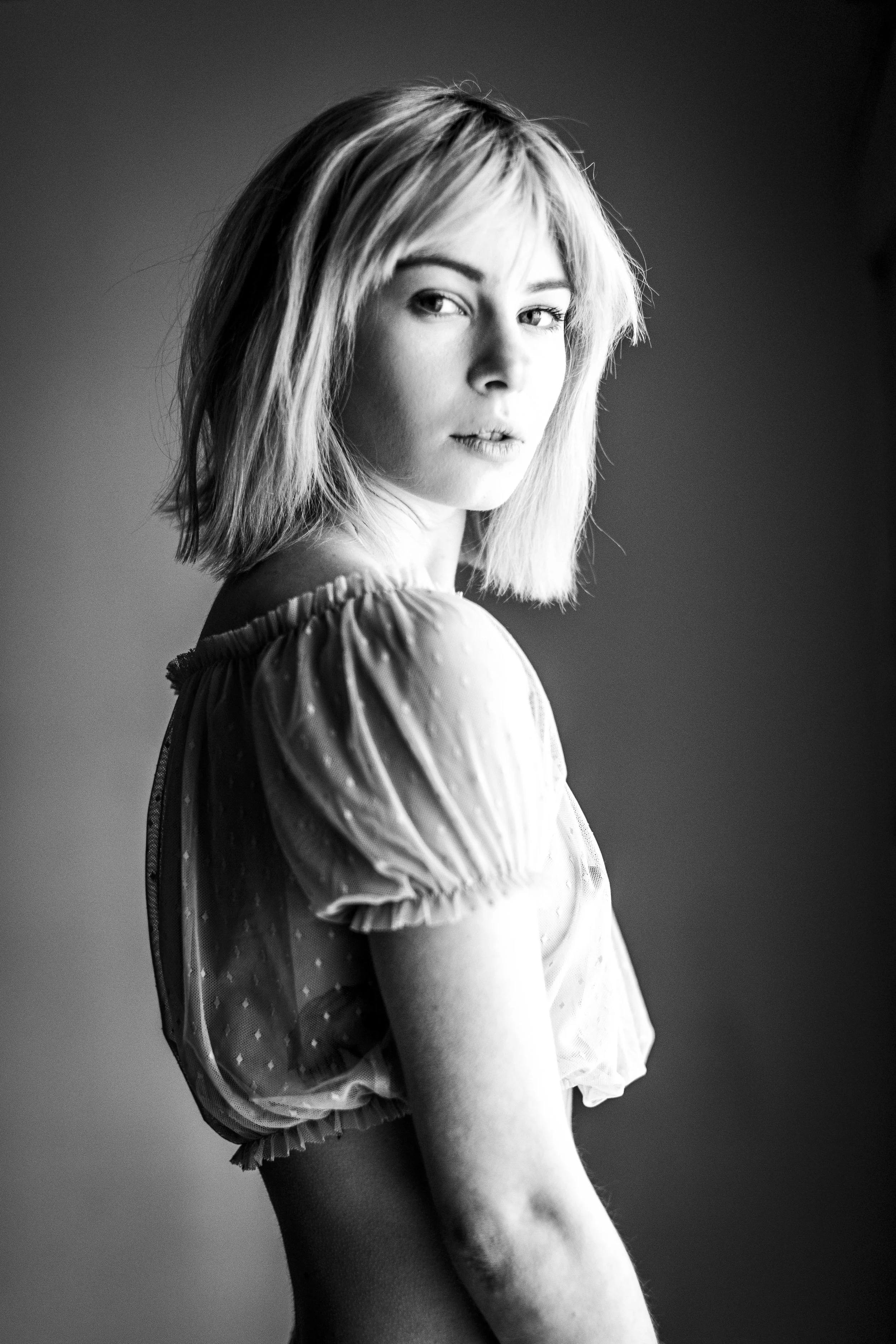 Black and white photo of a woman with shoulder-length hair, looking over her shoulder with a serious expression, wearing a puffed sleeve top.