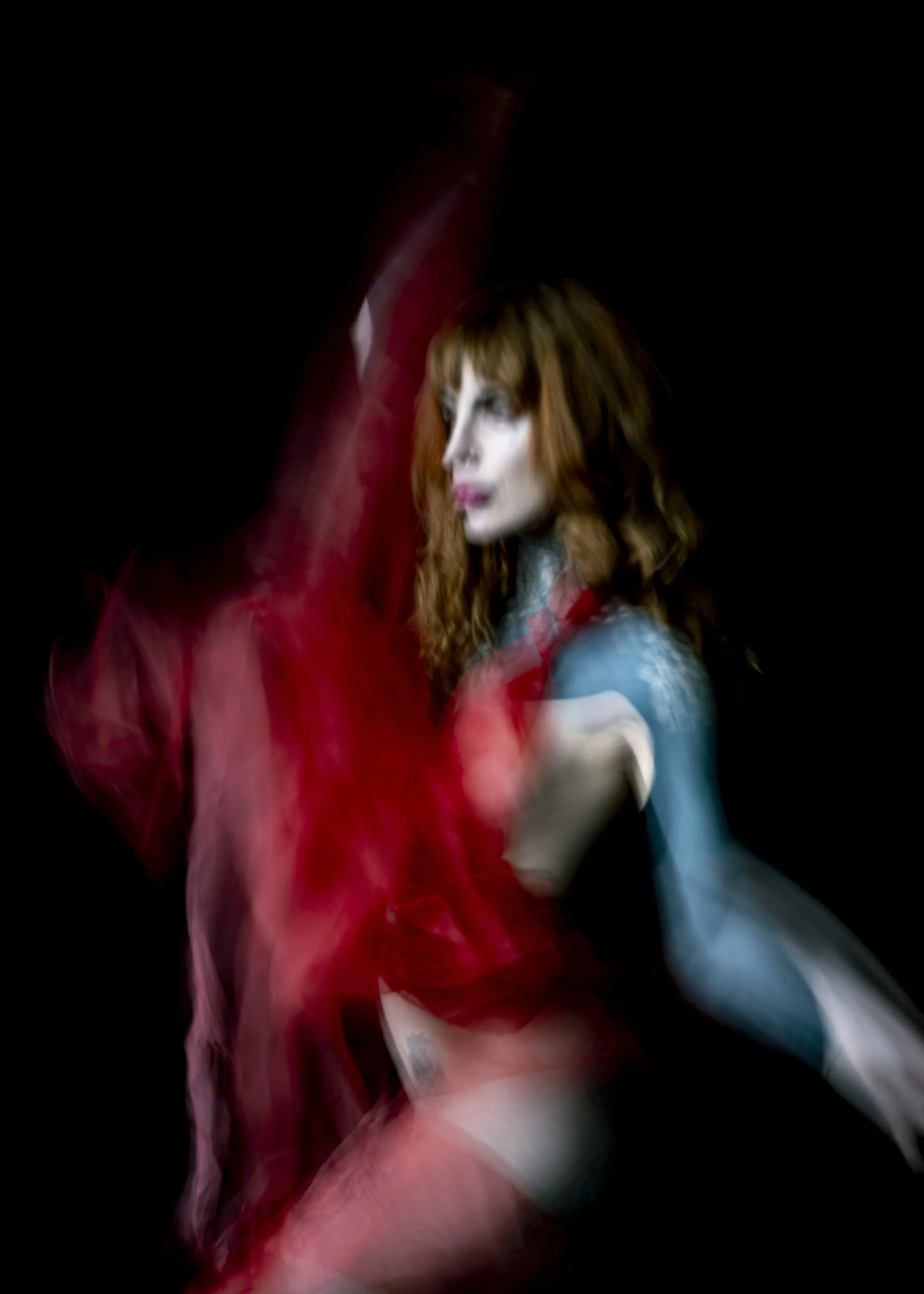 A woman with long red hair and bright makeup, wearing a blue top, posing against a black background with red sheer fabric flowing around her.