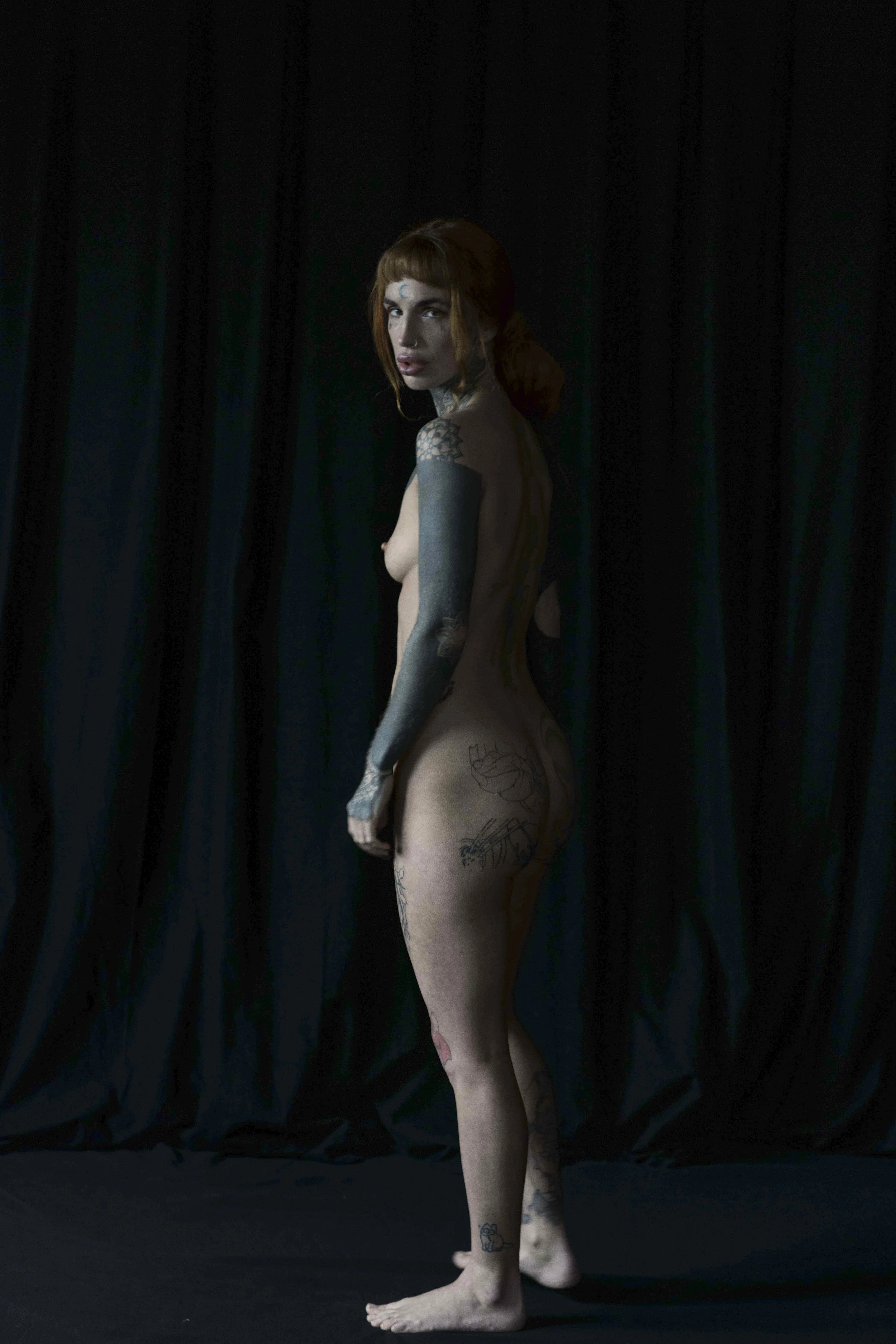 A young woman with tattoos, red hair, and blue eyes stands nude against a dark curtain background, looking over her shoulder.