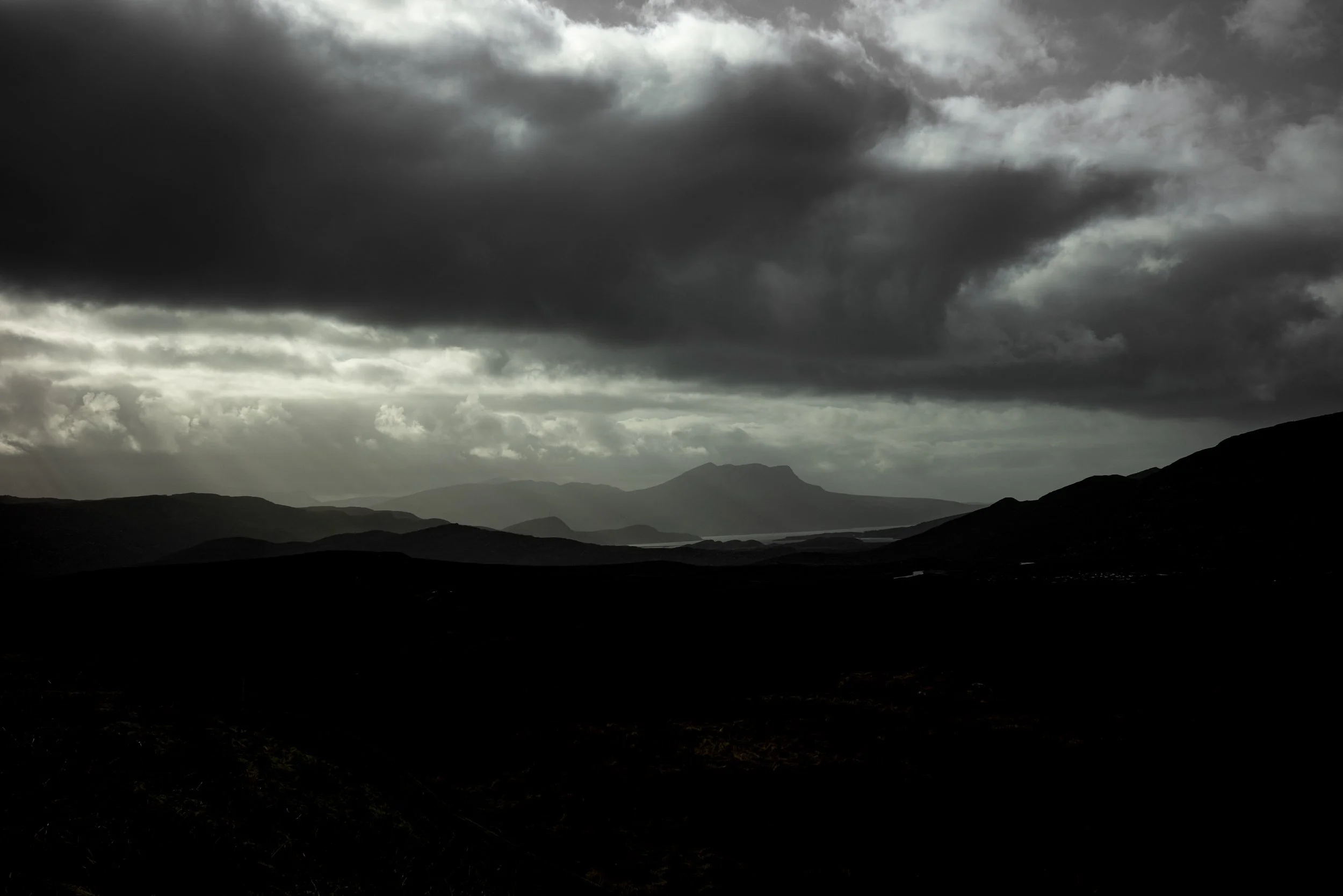 Dark storm clouds over a mountain landscape with hills and a river or lake in the distance.