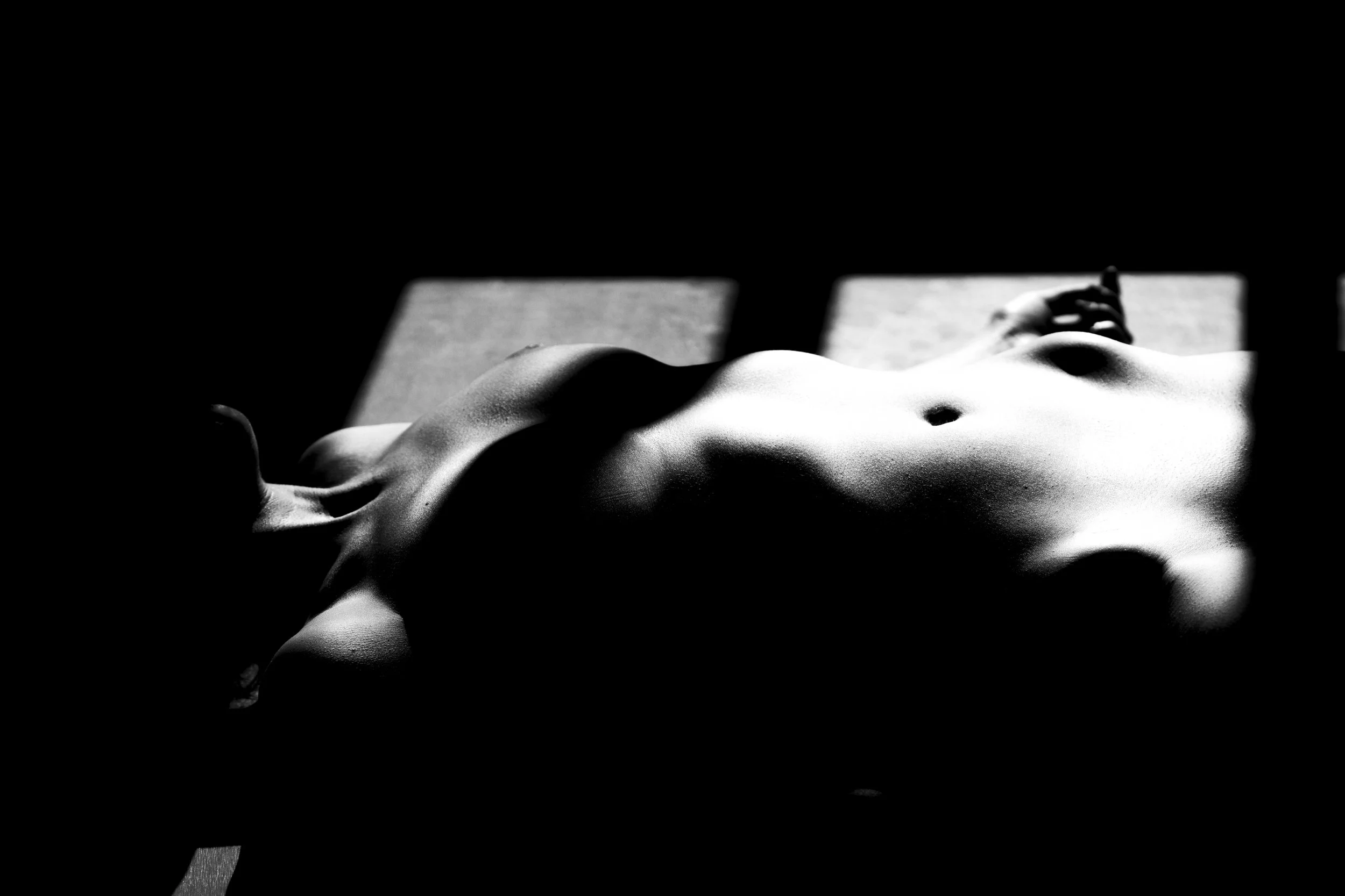 Black and white photo of a nude woman's body lying on a surface with shadows cast across her, with part of her face visible in the background.