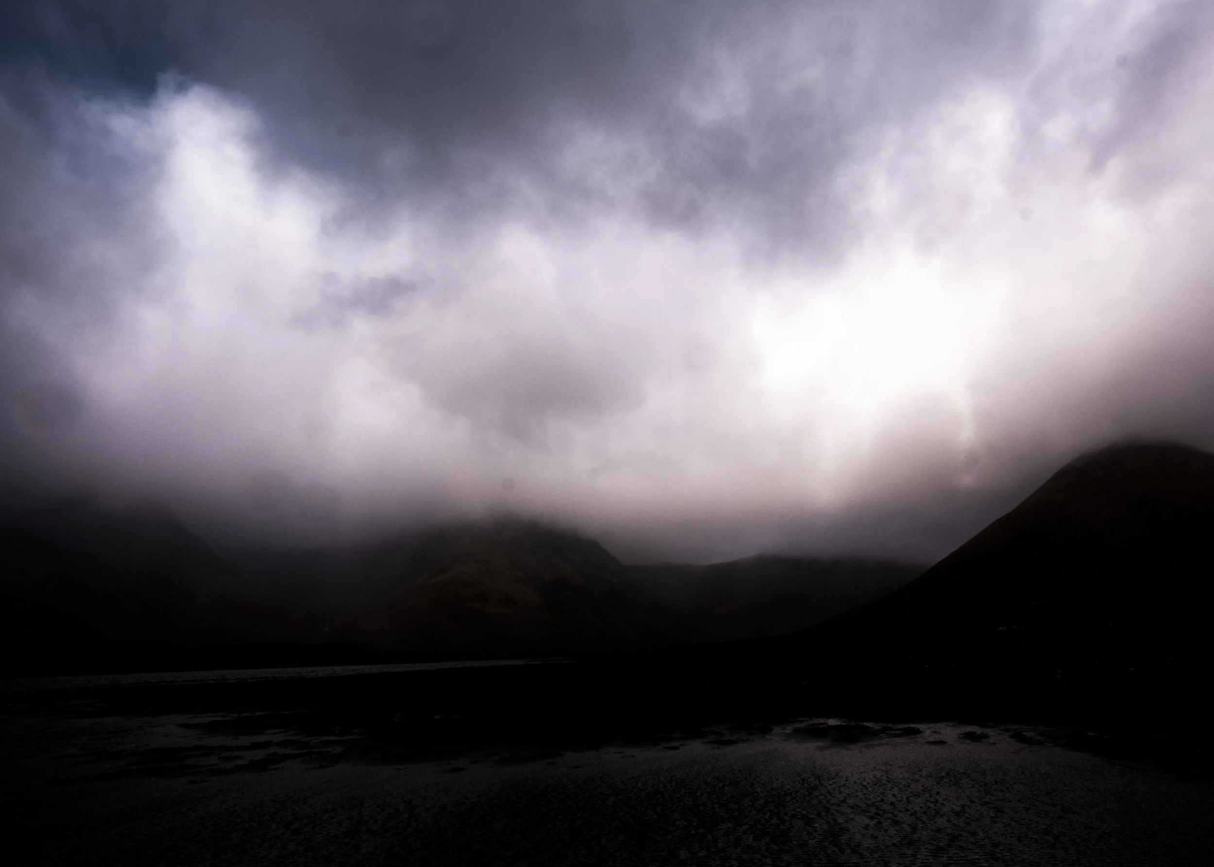 Dark stormy clouds over mountains with a body of water in the foreground.