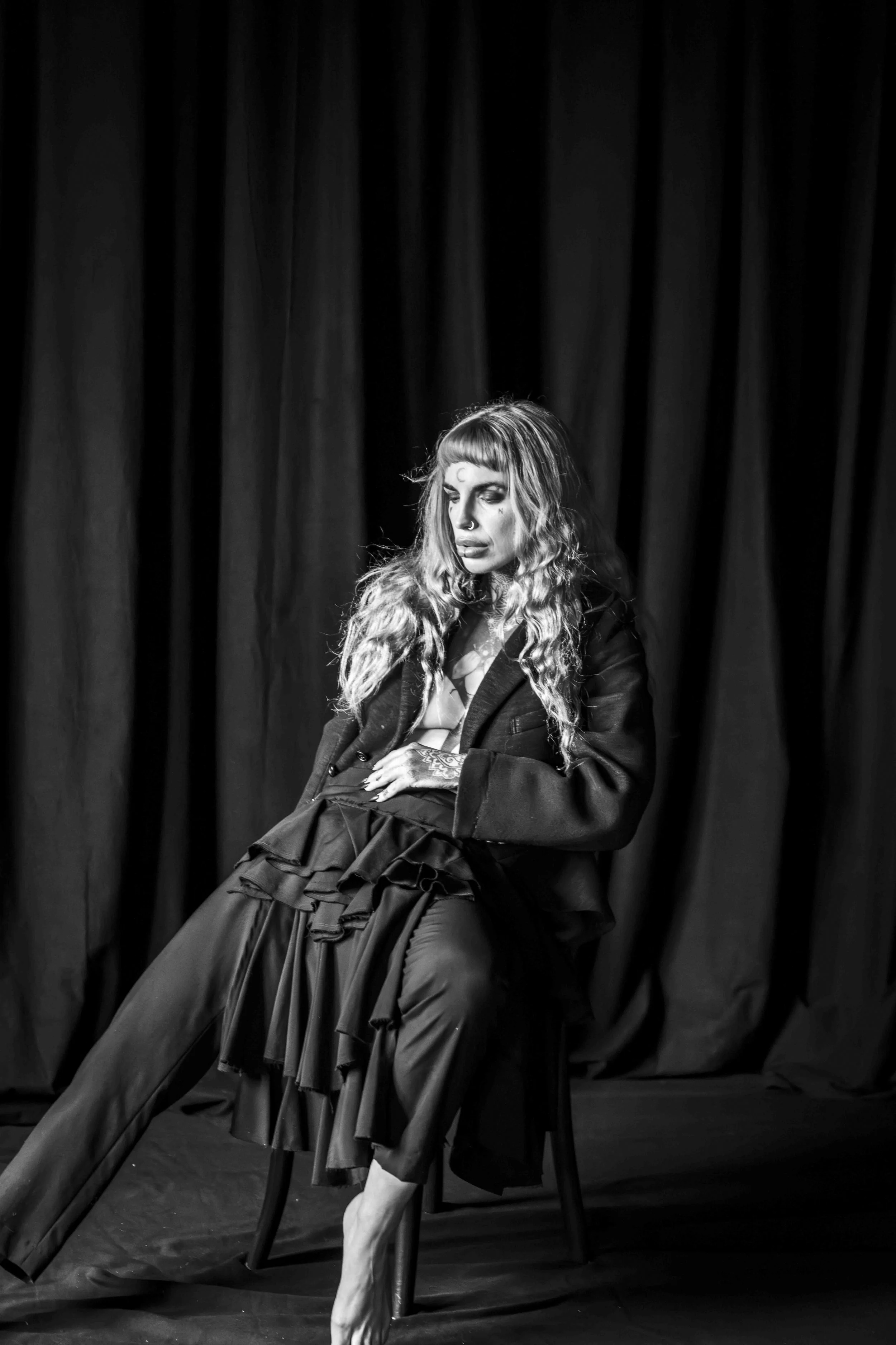 Black and white photo of a woman with long, curly hair and a septum piercing sitting on a chair with a dark curtain in the background. She has face tattoos and is wearing an oversized jacket. One leg is crossed over the other, and her foot is bare.