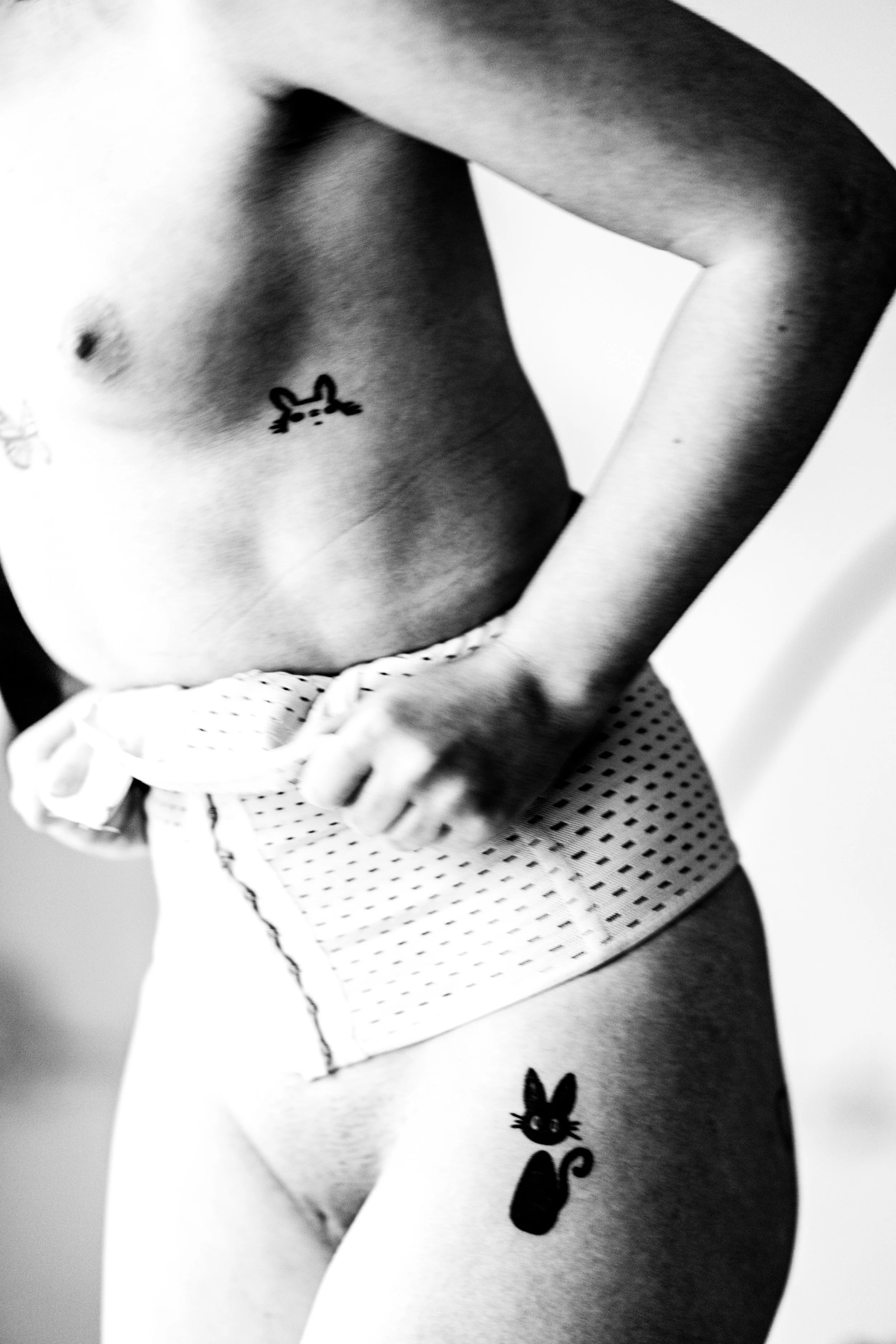 Close-up of a person's torso, showing a small bunny tattoo and a larger cat tattoo on their thigh. The person has the arm bent, with a hand resting on the waist.