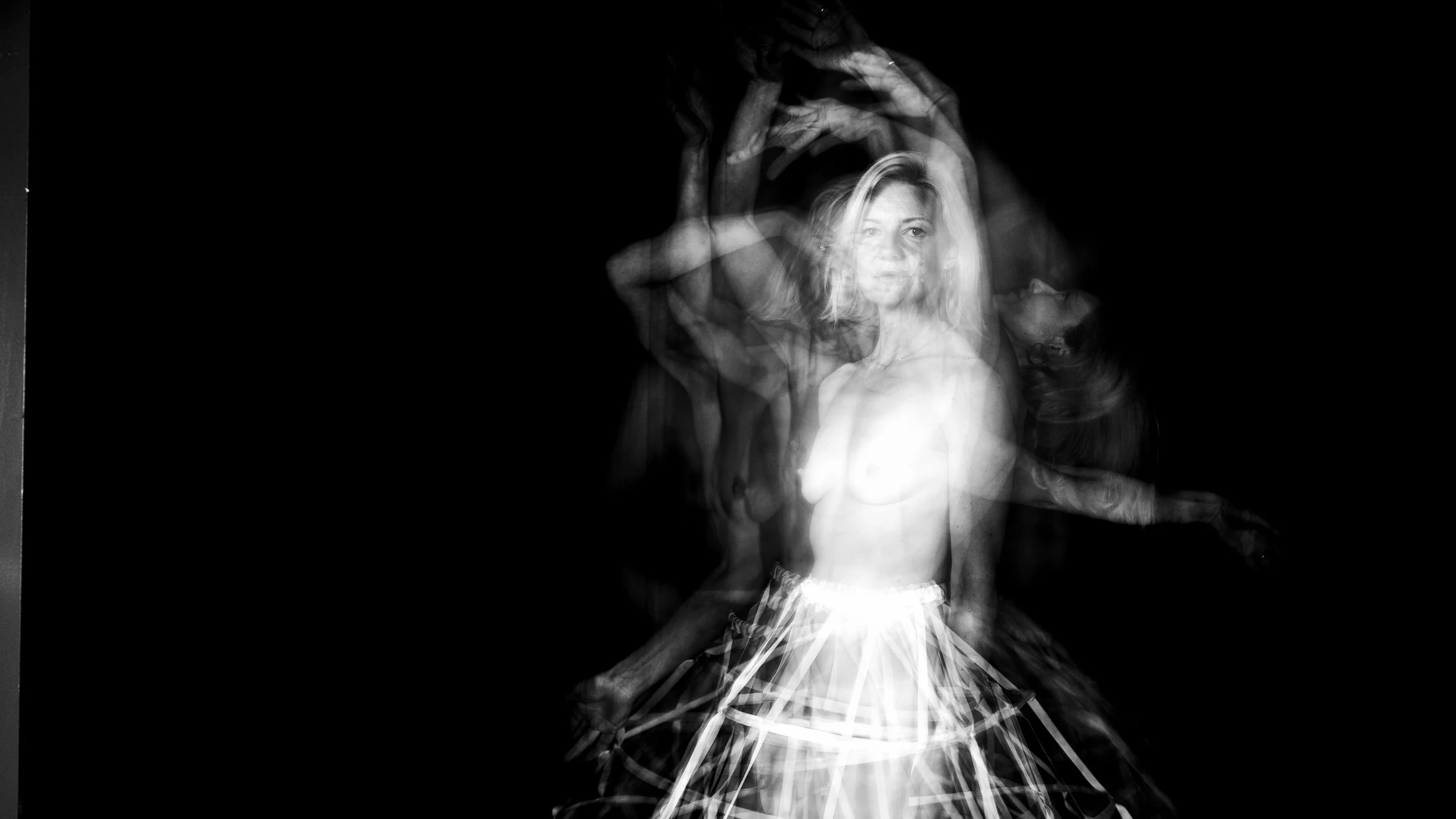 Black and white photo of a woman with light-colored hair, topless, wearing a skirt, surrounded by blurred, overlapping images of her moving arms and face.