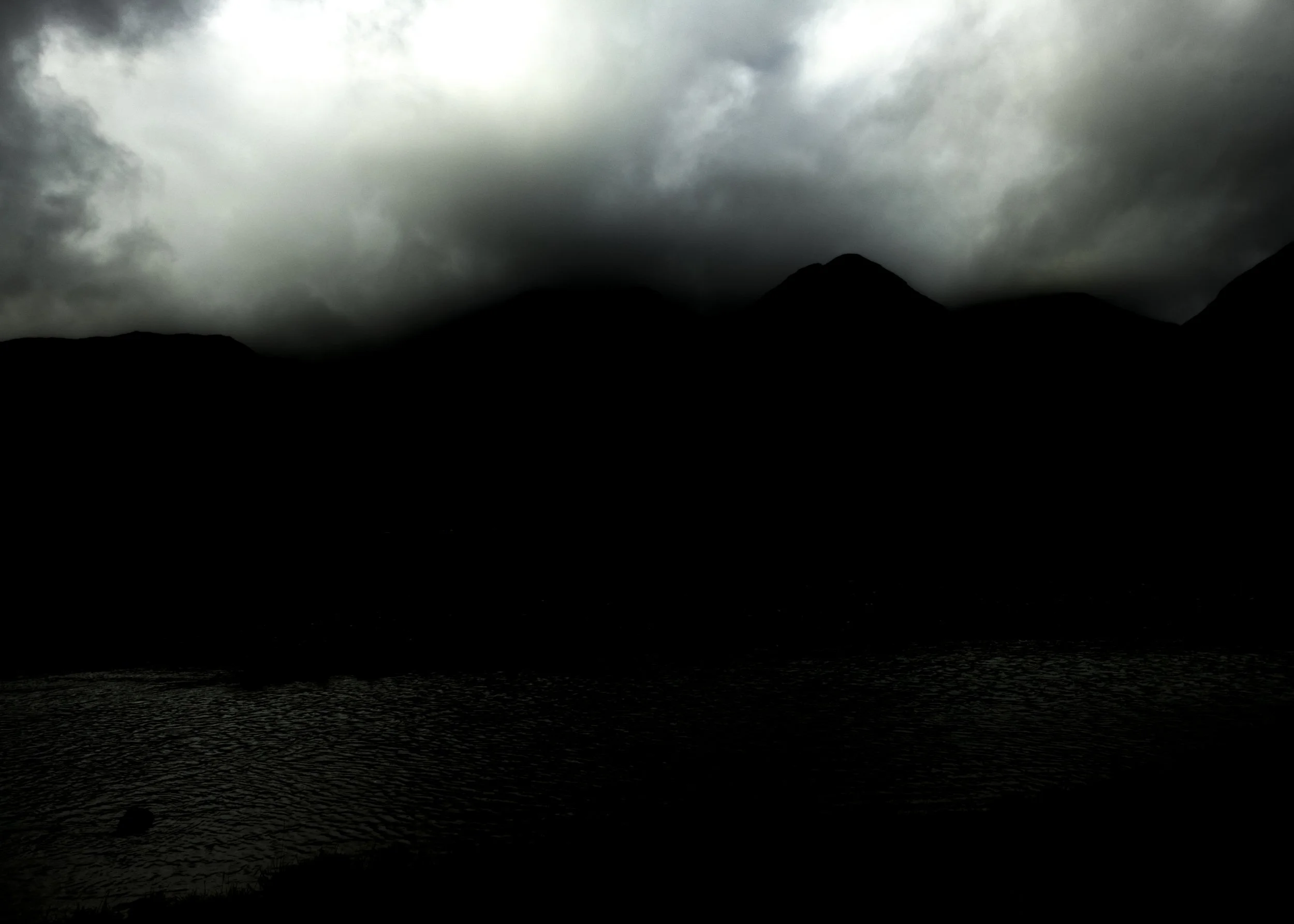 Dark stormy sky over a mountain silhouette and a body of water in the foreground.
