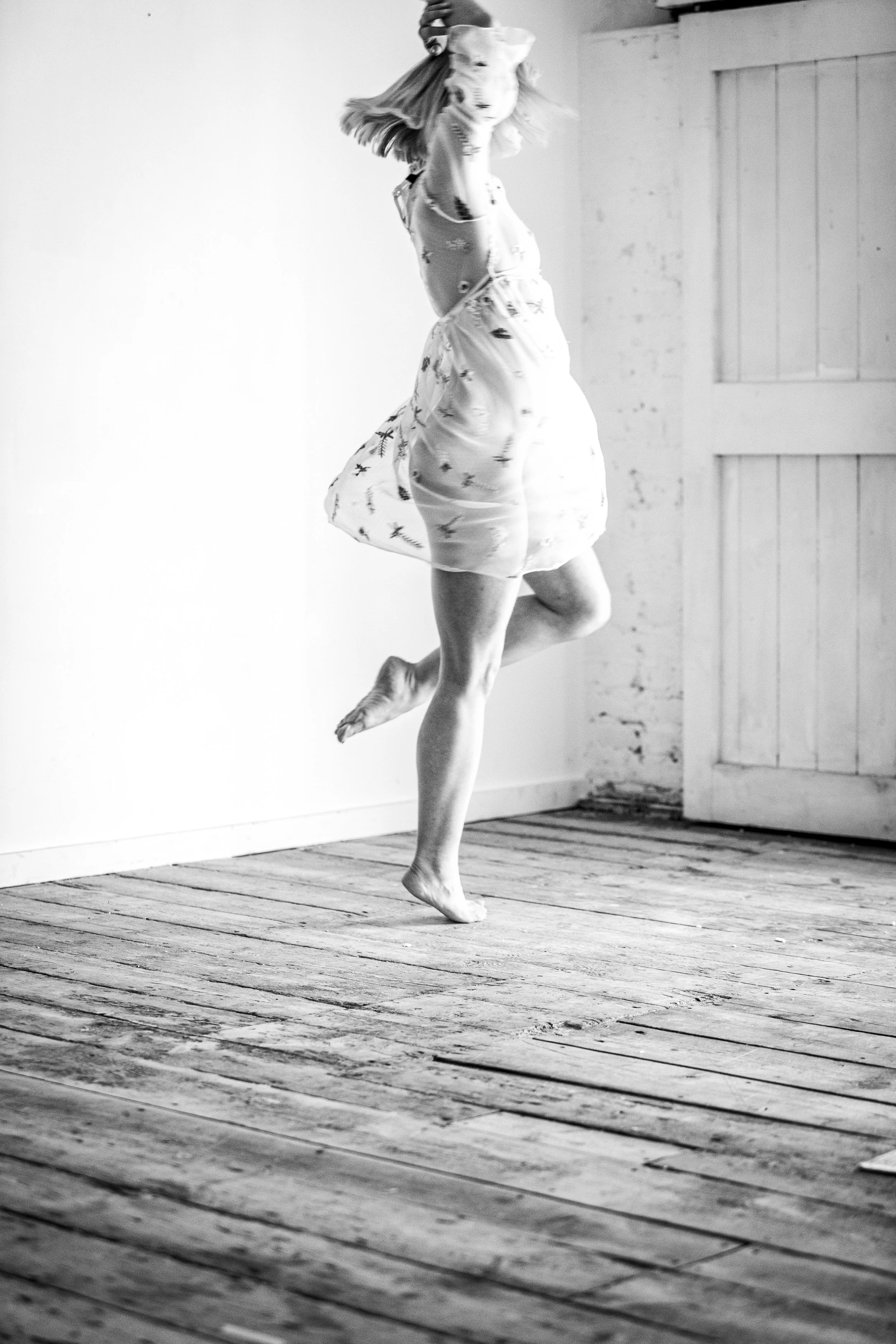 Black and white photo of a woman dancing or jumping on a wooden floor with an open door in the background.