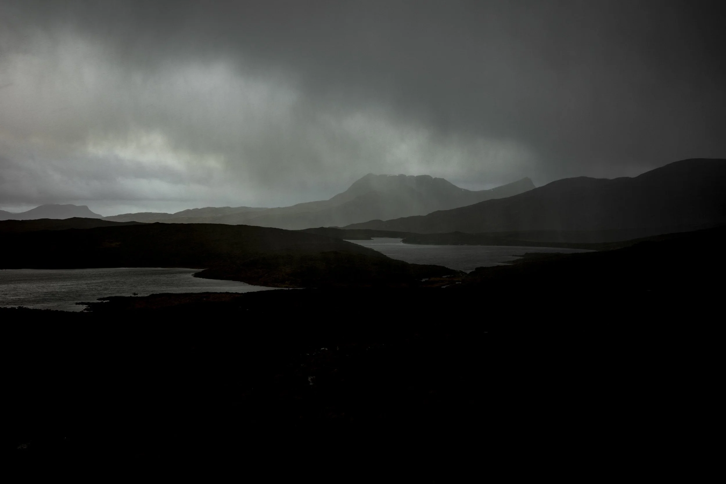 Dark stormy skies over a mountainous landscape with water and rolling hills in the foreground.