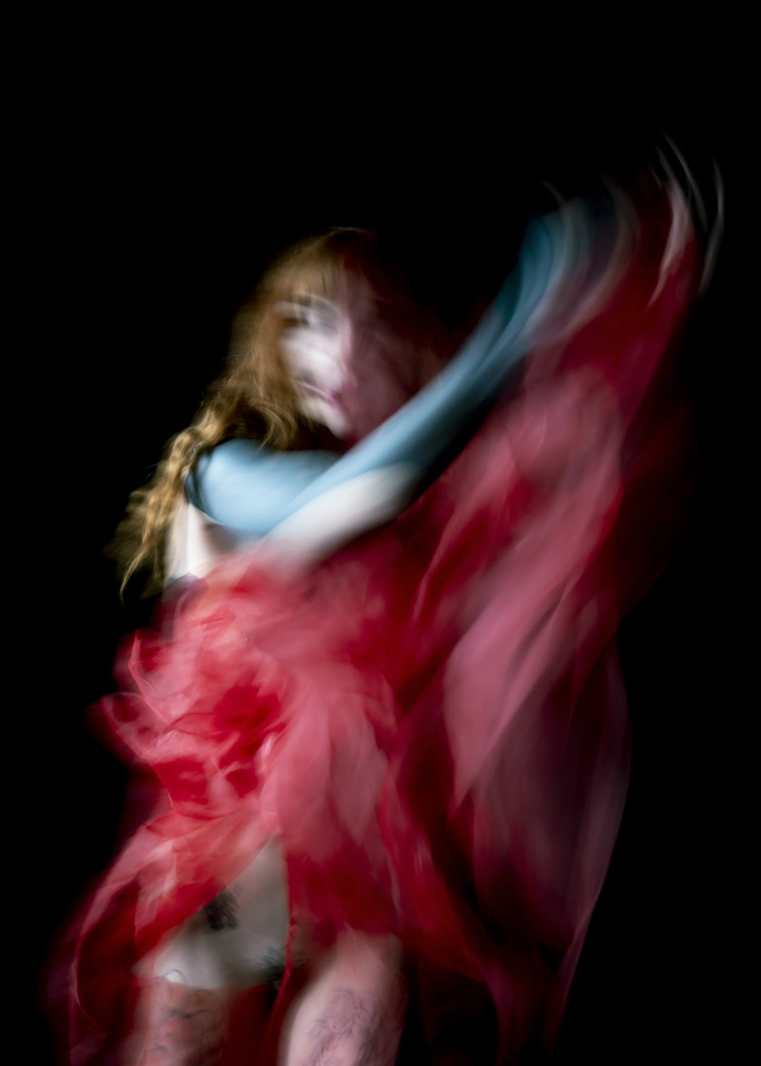 Blurred image of a woman with long curly hair, wearing colorful fabric, against a black background.