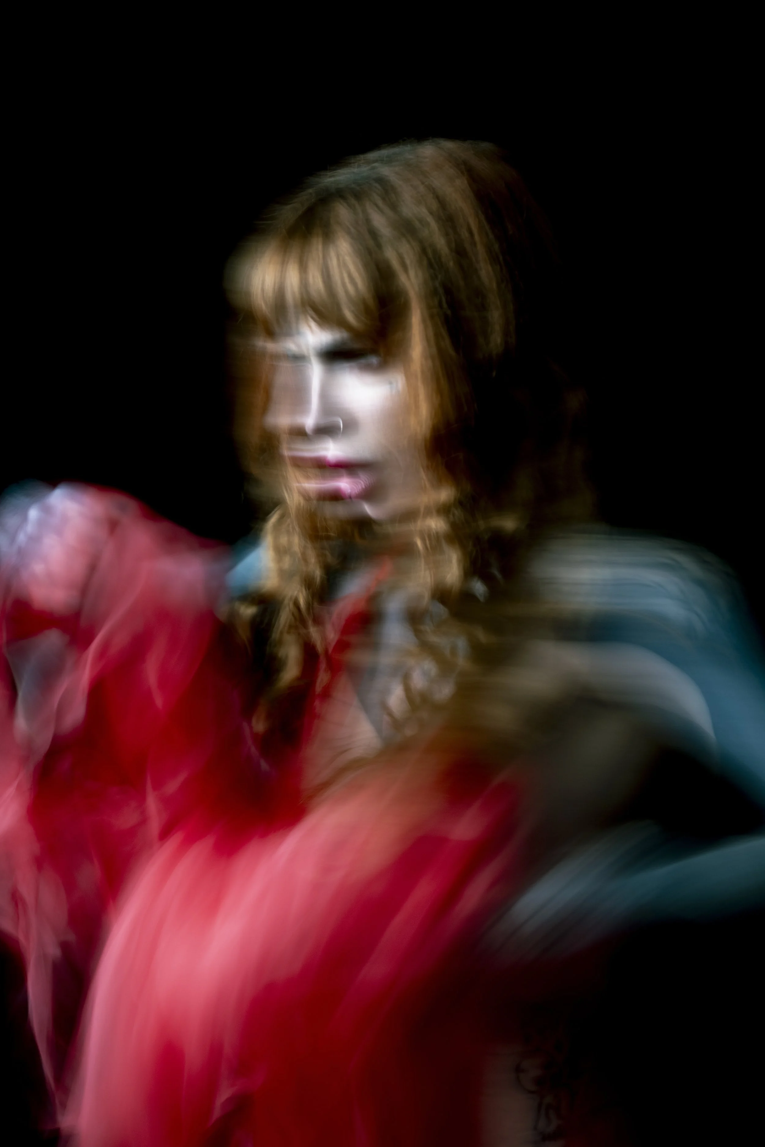 A woman with long, wavy, reddish-brown hair and bangs, wearing a red top, with a serious expression. The image appears to have motion blur against a black background.