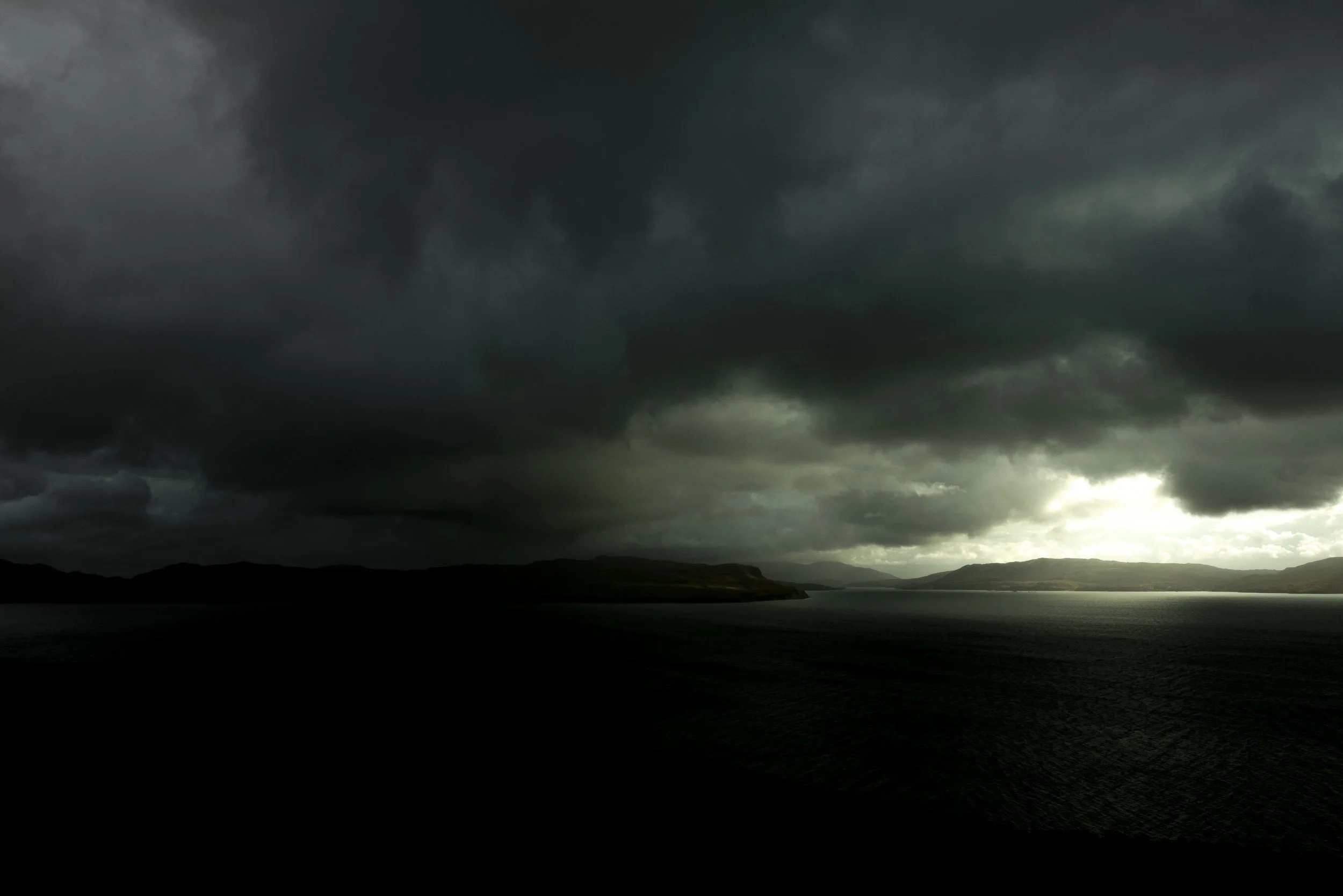 Dark, stormy clouds over a landscape with water and distant hills.