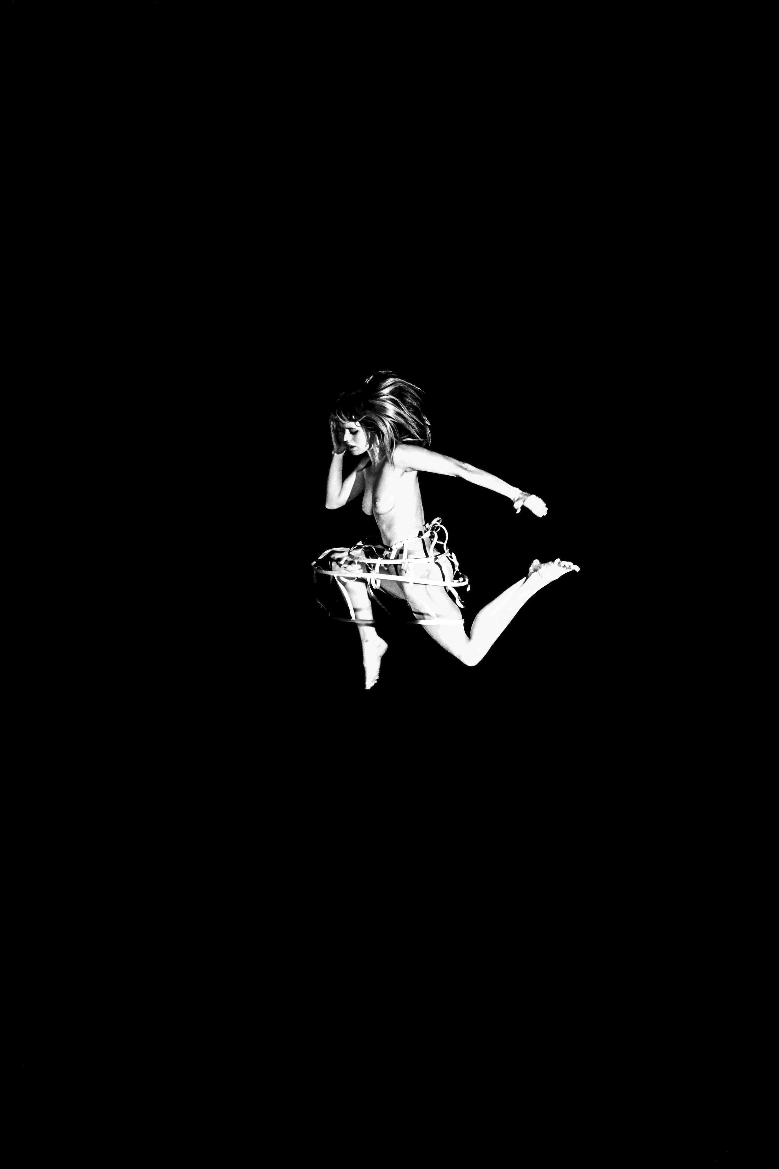 A woman with long hair, topless, with her hand on her head, sitting inside a transparent plastic hula hoop, against a black background.