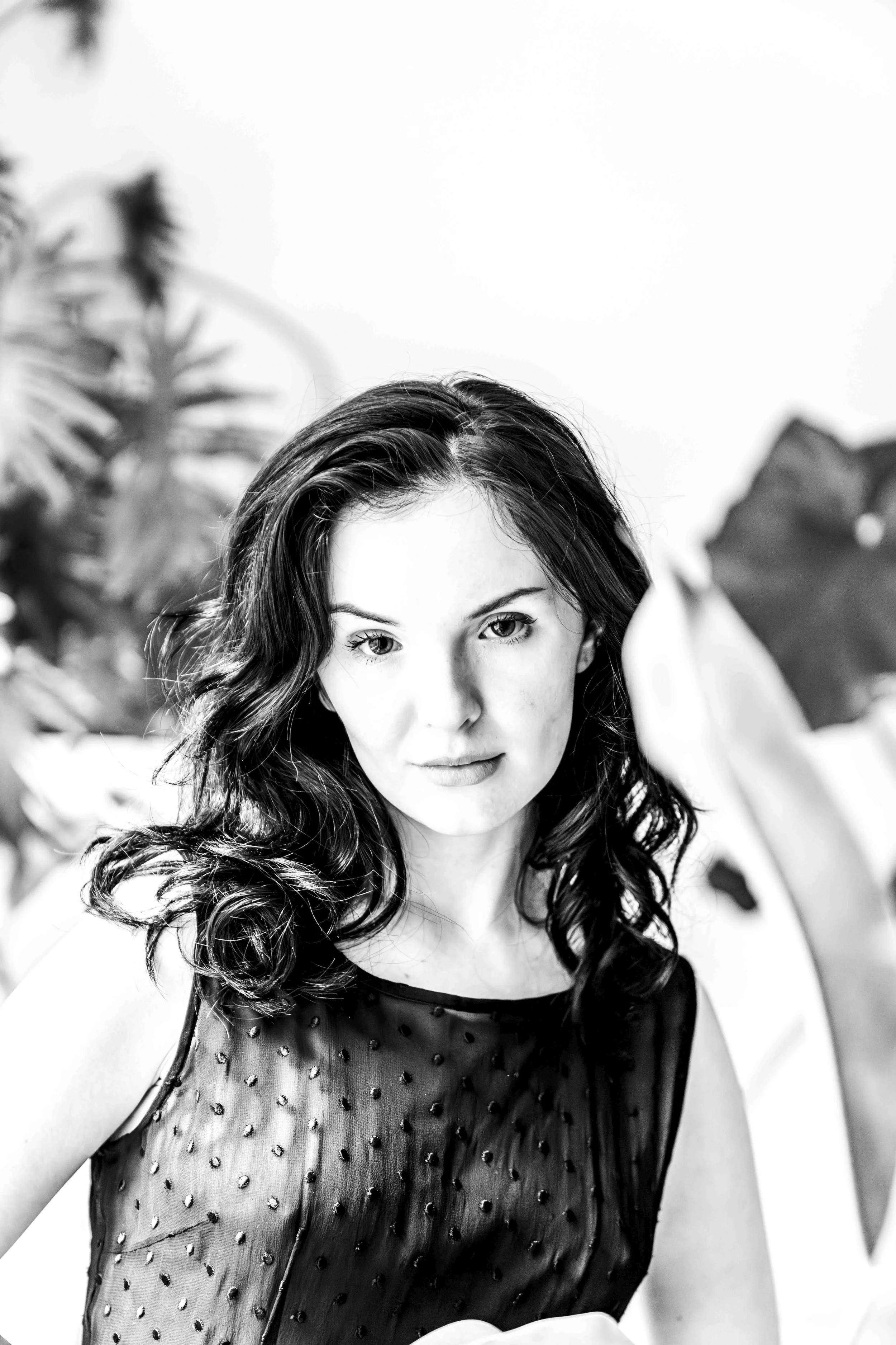 A black and white photo of a woman with wavy hair, looking directly at the camera, with a large leafy plant in the background.