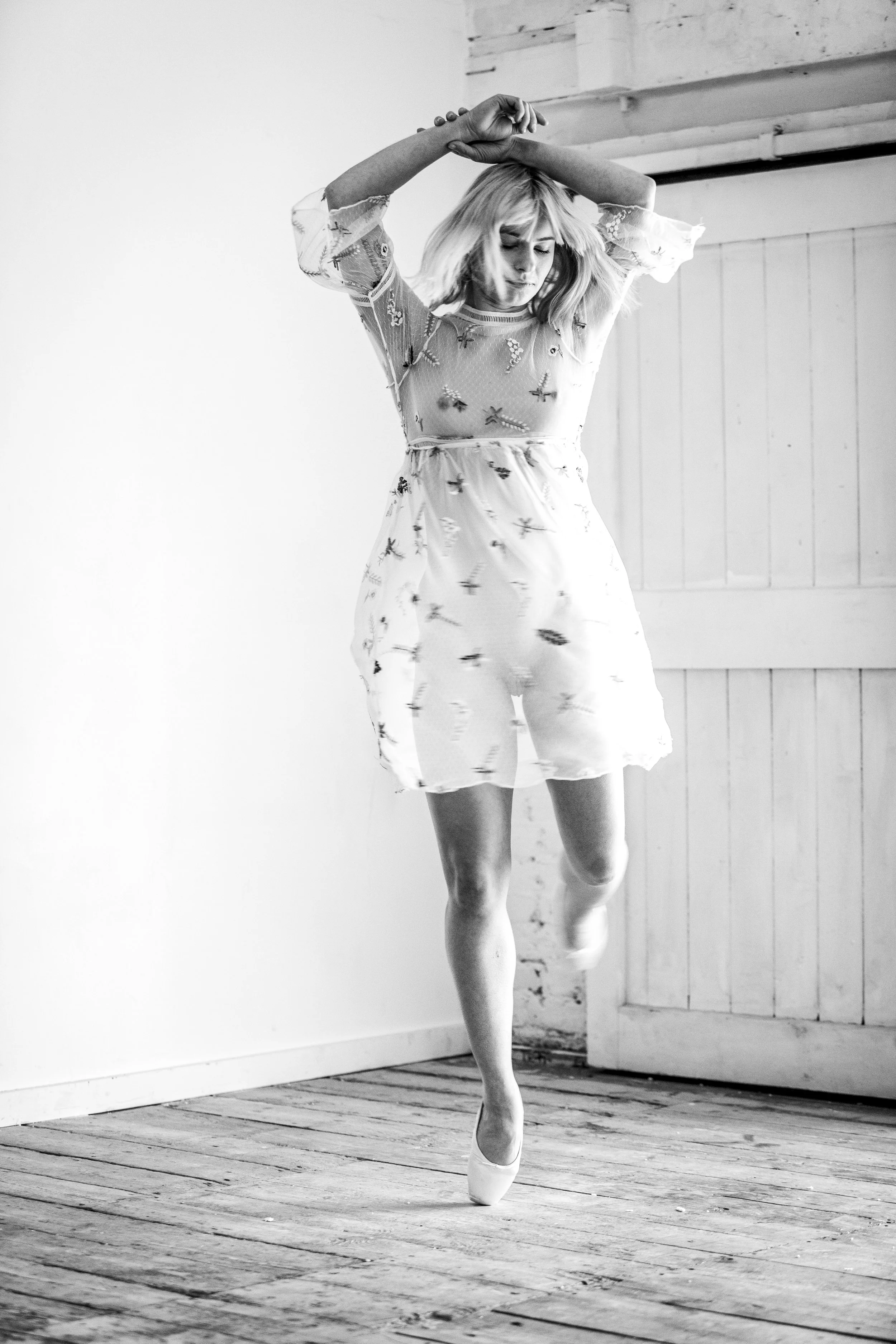 A woman with blonde hair in a dress with embroidered designs balances on one foot with arms crossed above her head in a dance pose indoors.