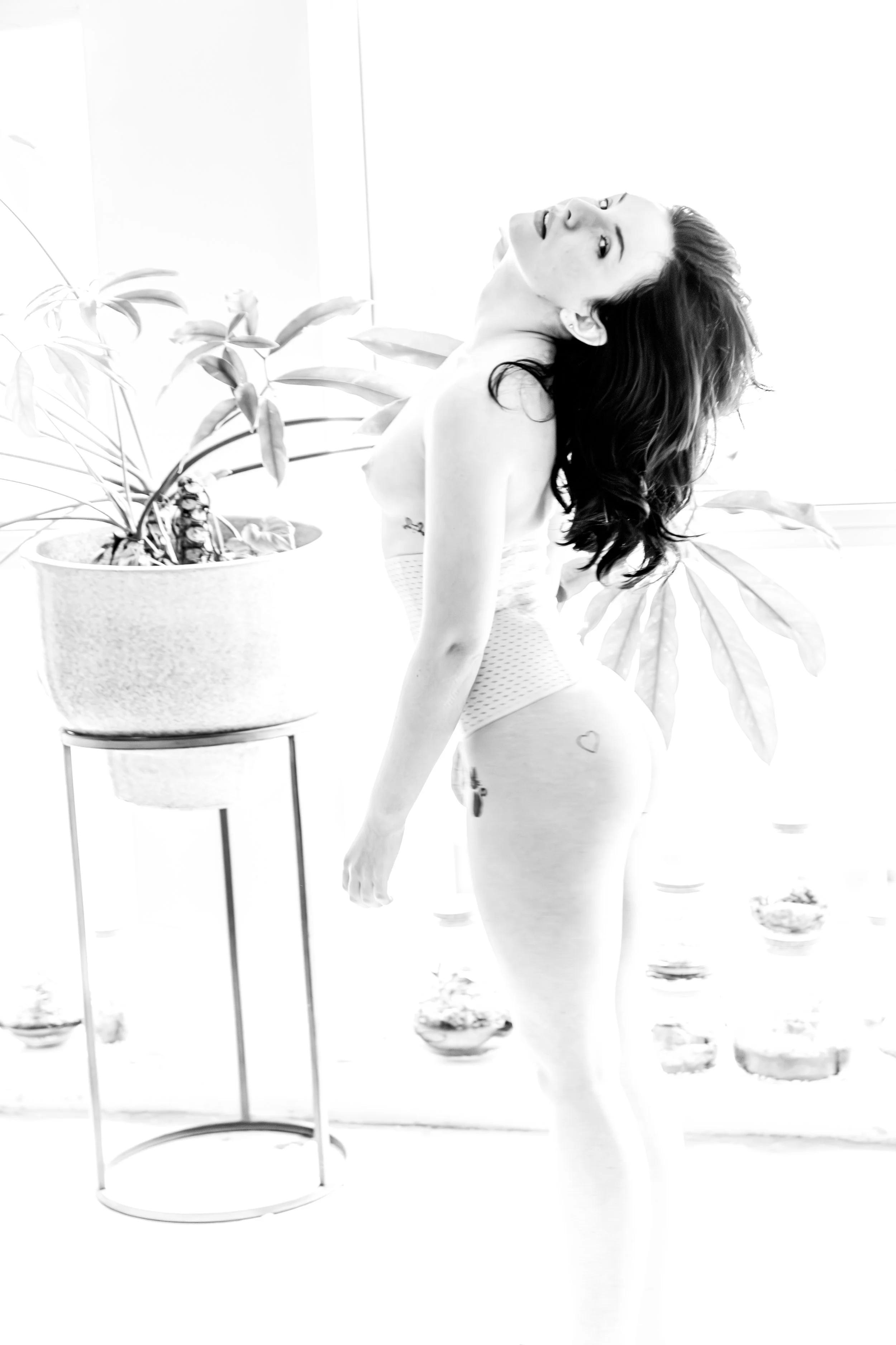A woman with long dark hair and tattoos on her side, wearing underwear, is standing by a potted plant with large leaves. The background appears bright and minimalist.