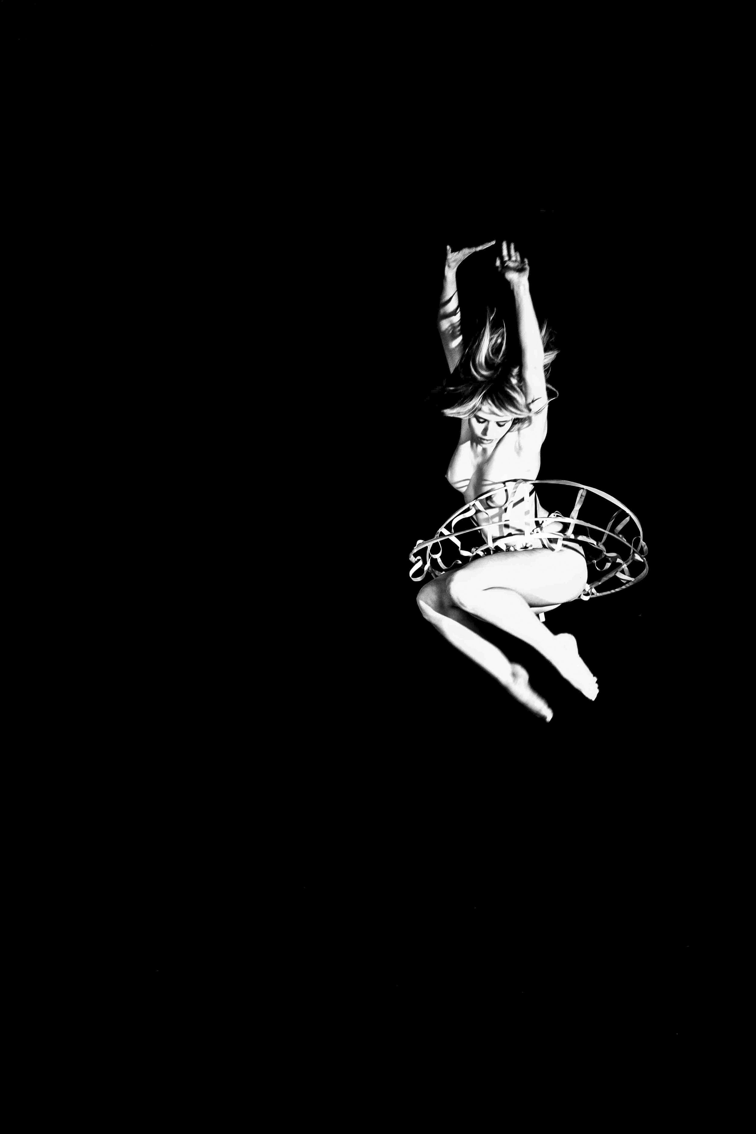 Black and white photograph of a female dancer mid-performance, wearing a tutu, with her arms raised and hair flowing, against a dark background.