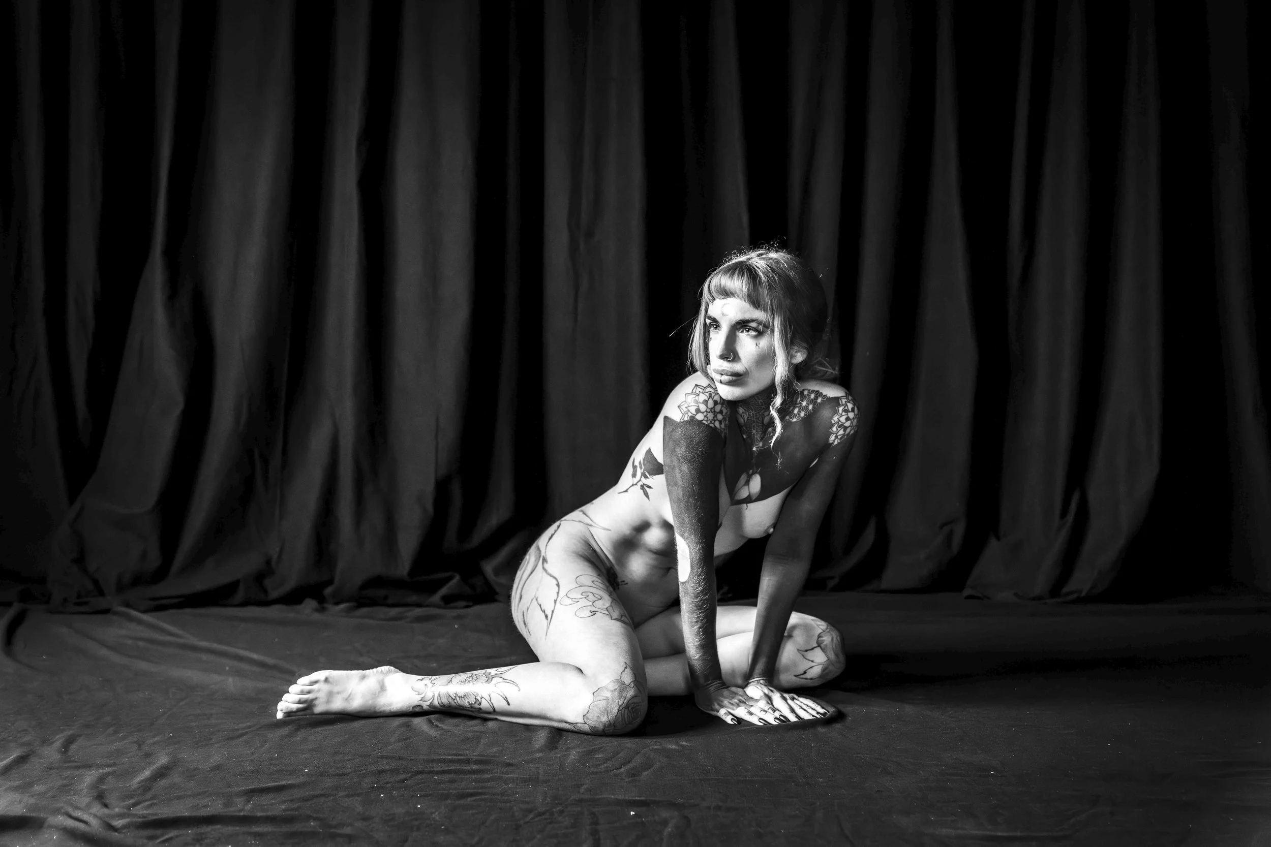 Black and white photo of a woman with tattoos sitting on the floor in front of dark curtains. She is on her hands and knees, looking to the right with an intense expression.