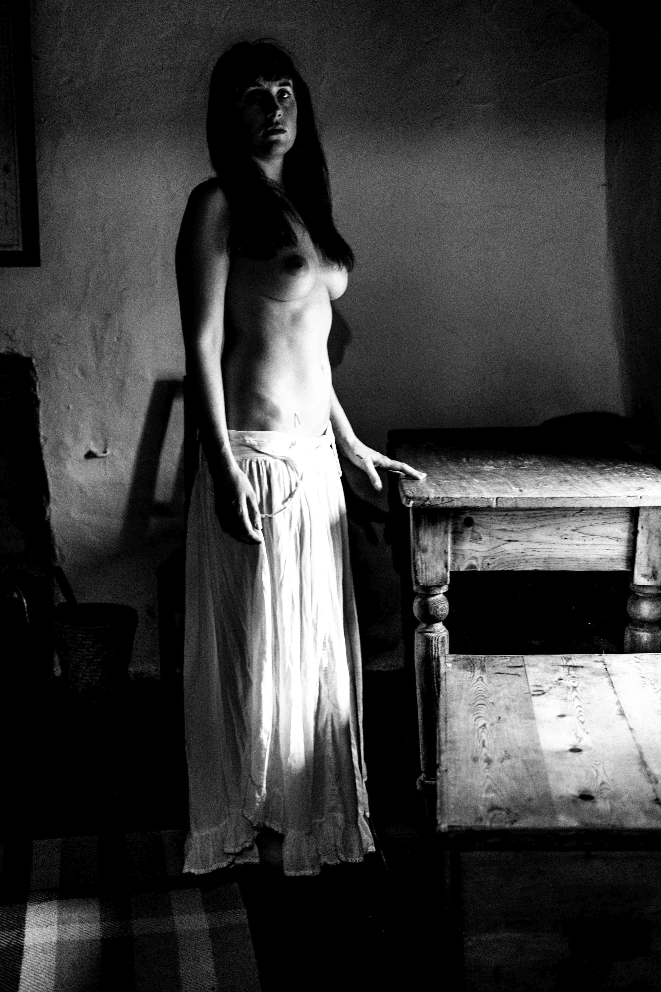 A black and white photo of a woman with long hair and bangs, standing in a room with textured walls. She is topless, wearing a long, flowing skirt, and resting her hand on a wooden table. The room has dim lighting, and the woman is looking directly at the camera with a serious expression.