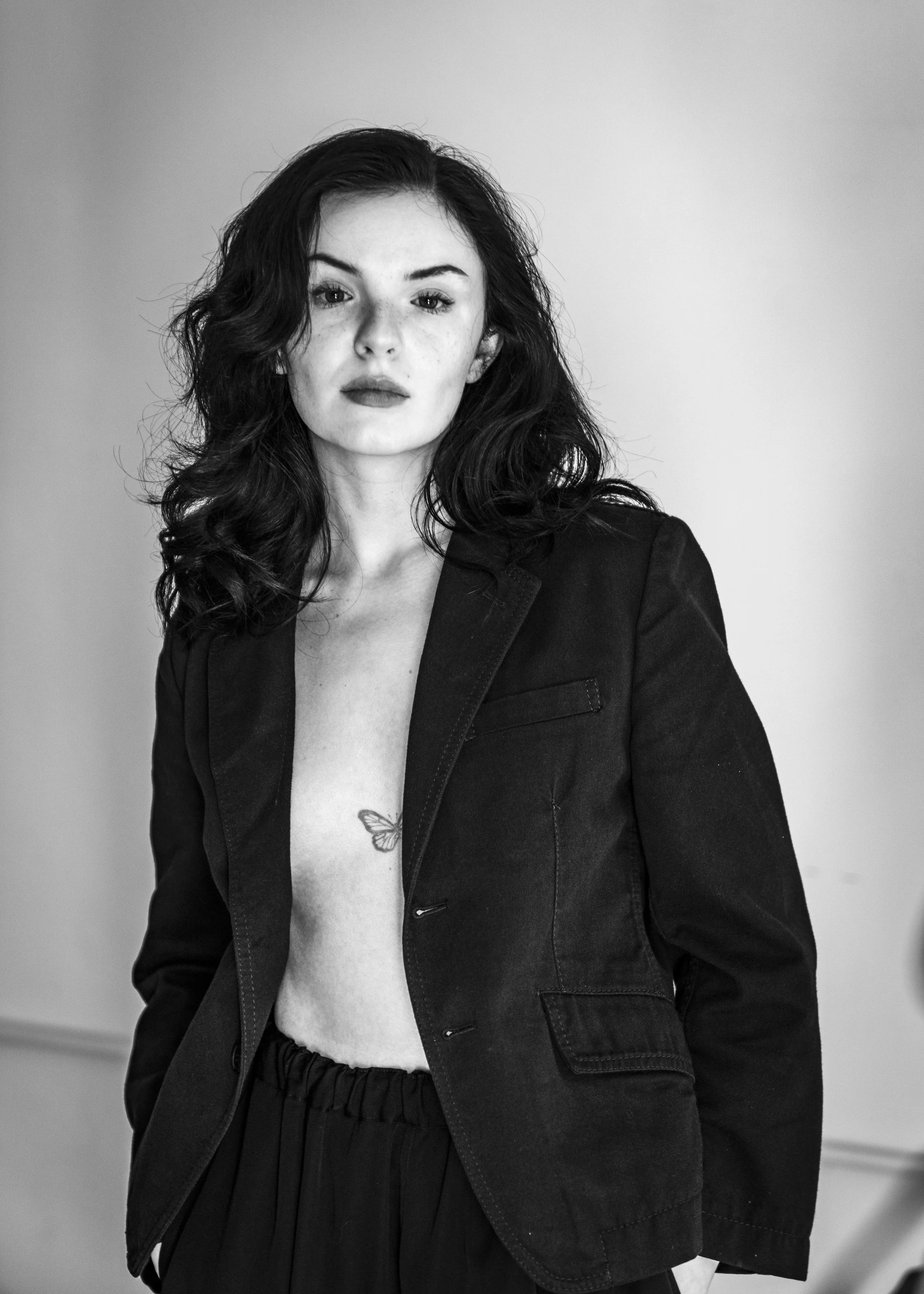A black and white photo of a woman with dark hair, wearing a blazer with no shirt underneath, revealing a butterfly tattoo on her chest. She is looking straight at the camera with a confident expression.