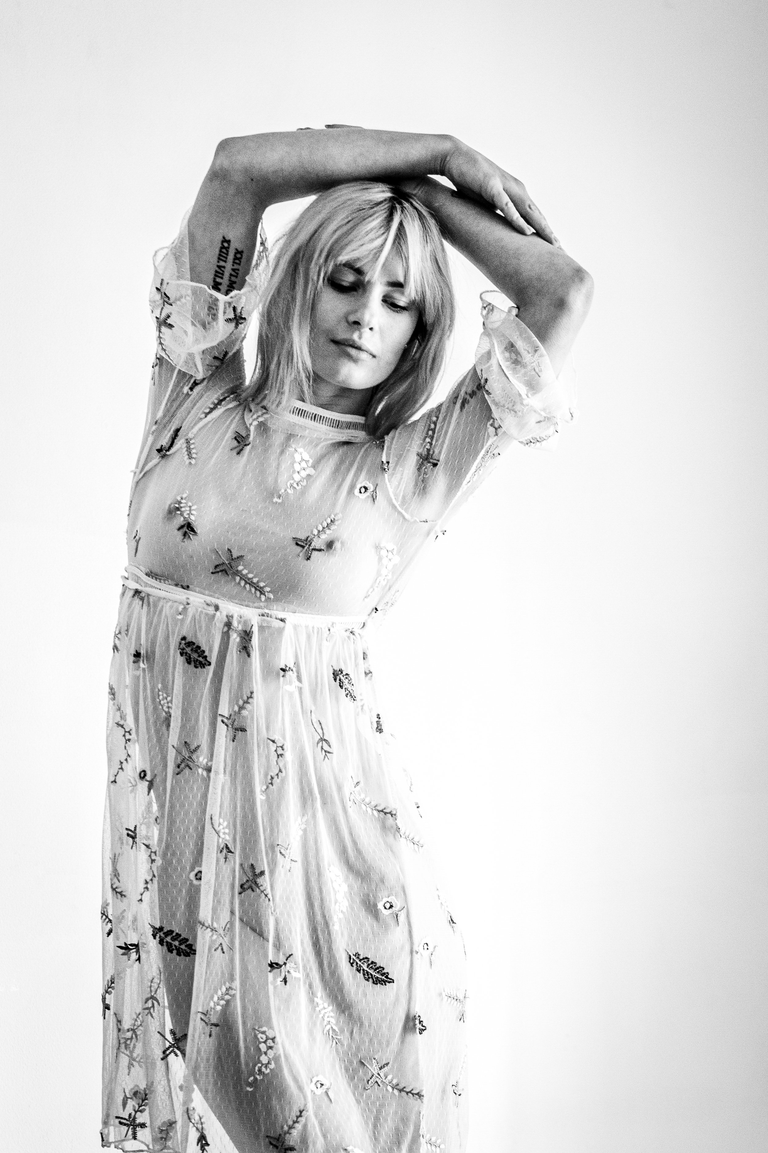 A woman with blonde hair with bangs, dressed in a sheer, embroidered dress with floral patterns, poses with her arms stretched above her head against a plain white background.