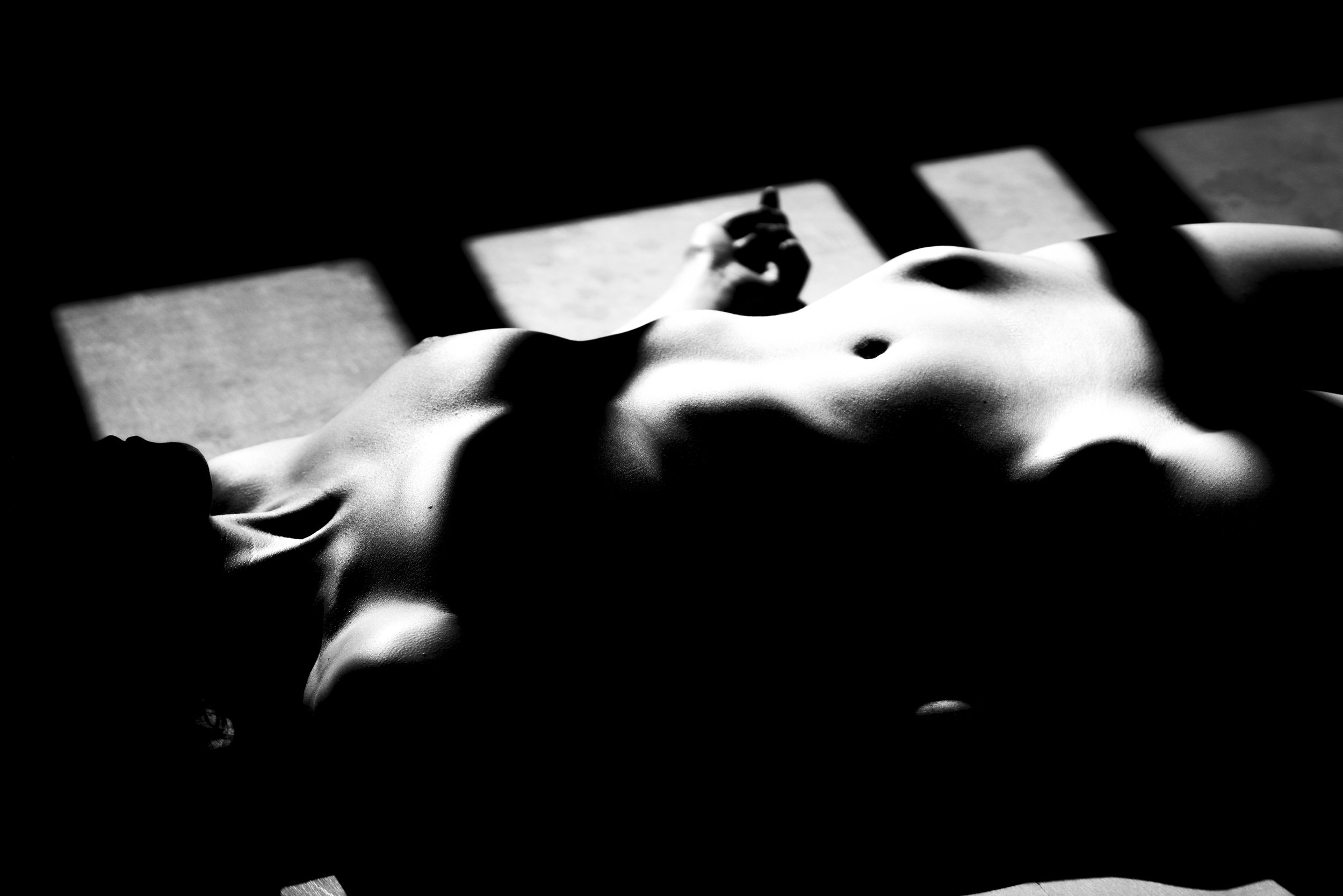 Black and white photo of a nude woman lying down, with shadows cast across her body by a grid or bars.