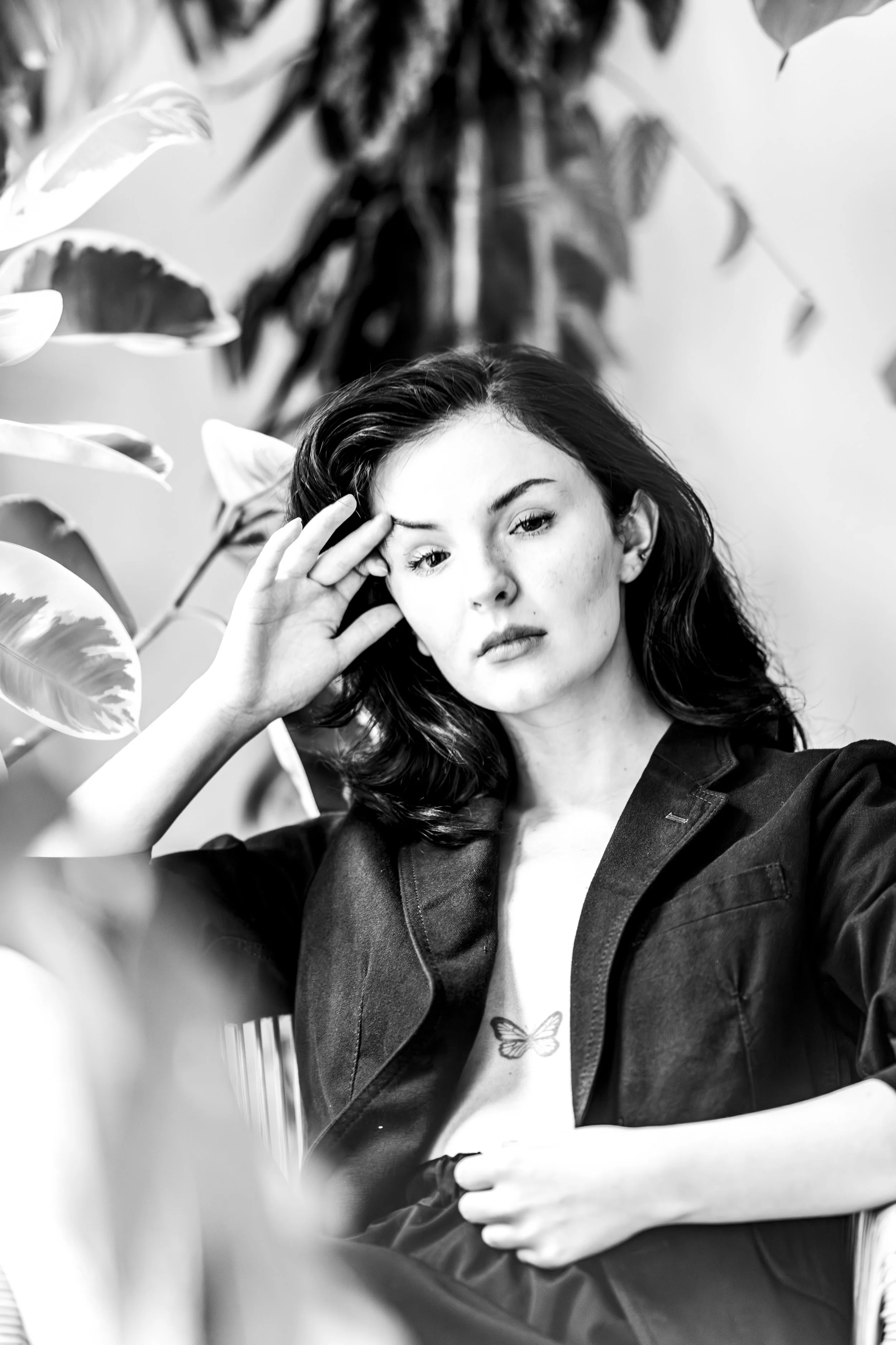 Black and white photo of a woman with dark hair sitting among large indoor plants, with her hand resting on her forehead, wearing a dark jacket and a T-shirt with a butterfly print.