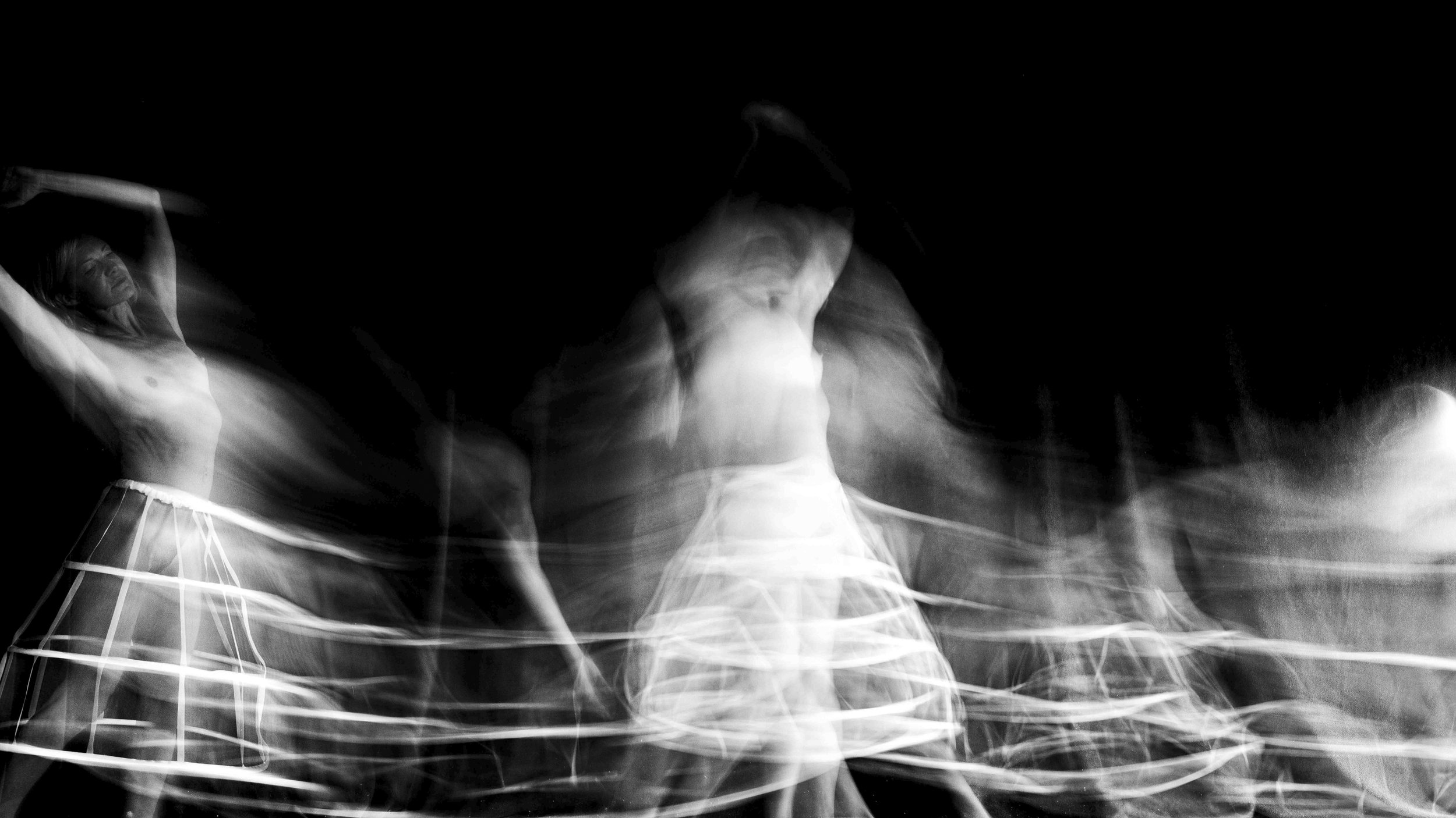 Black and white photograph of a woman dancing, captured with motion blur creating a sense of movement around her, wearing a striped skirt and a sleeveless top.