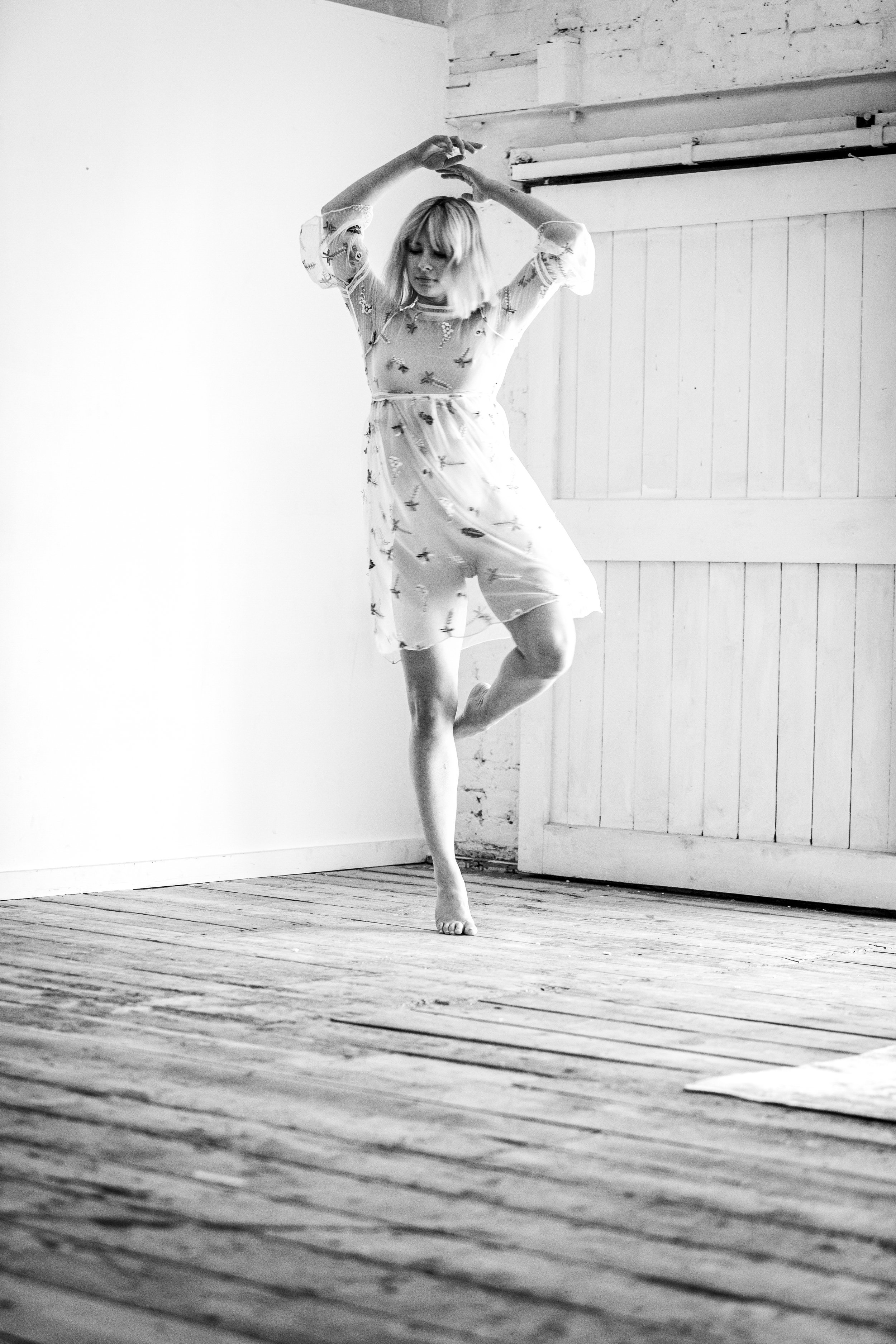 Black and white photo of a woman dancing barefoot on a wooden floor in a rustic room. She's wearing a light, semi-transparent dress with embroidered details, and has short, blonde hair. Her arms are raised above her head as she strikes a dance pose.