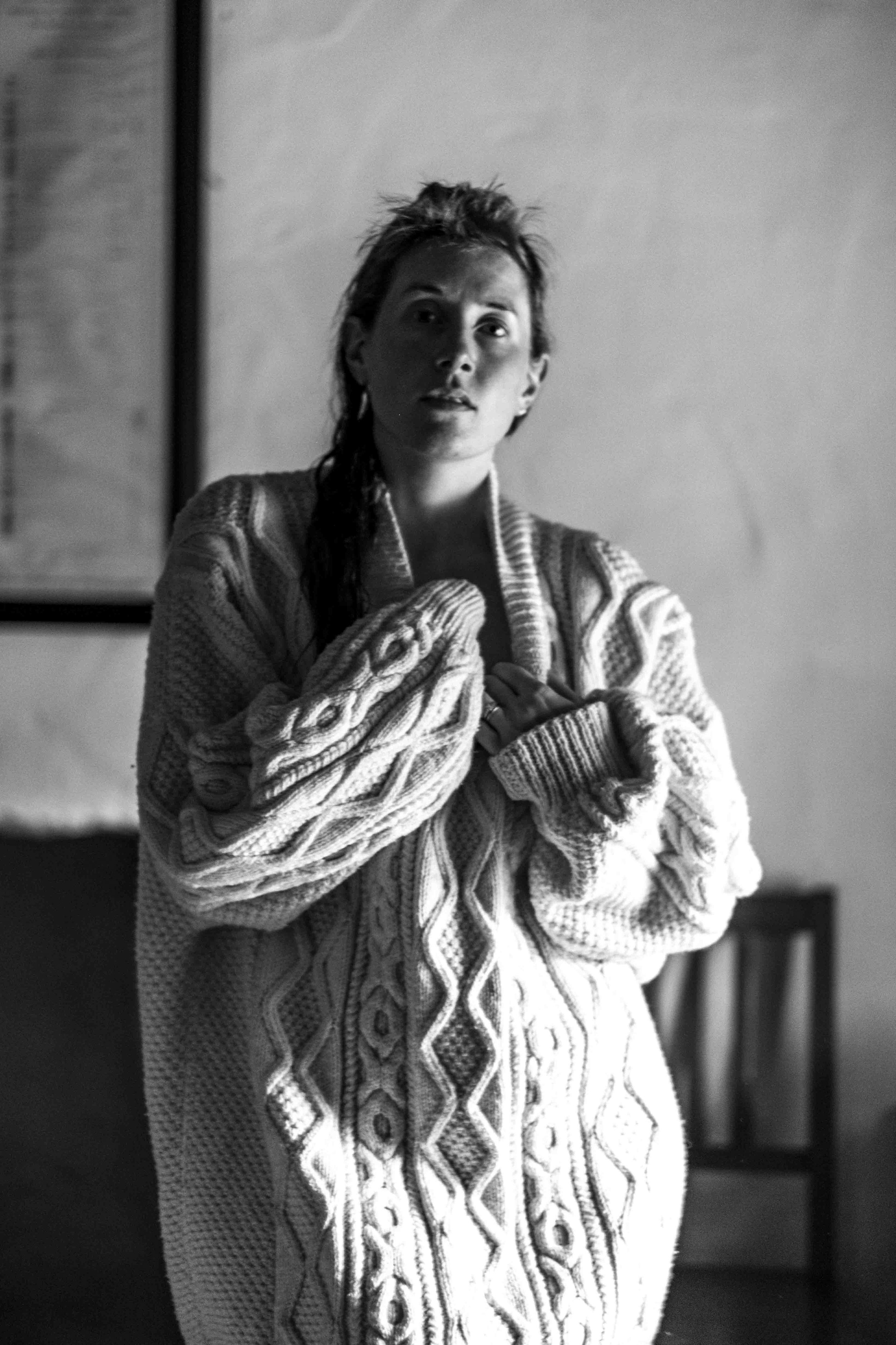 Black and white photo of a person with short, messy hair wearing a thick, patterned sweater, standing indoors with blurred background.