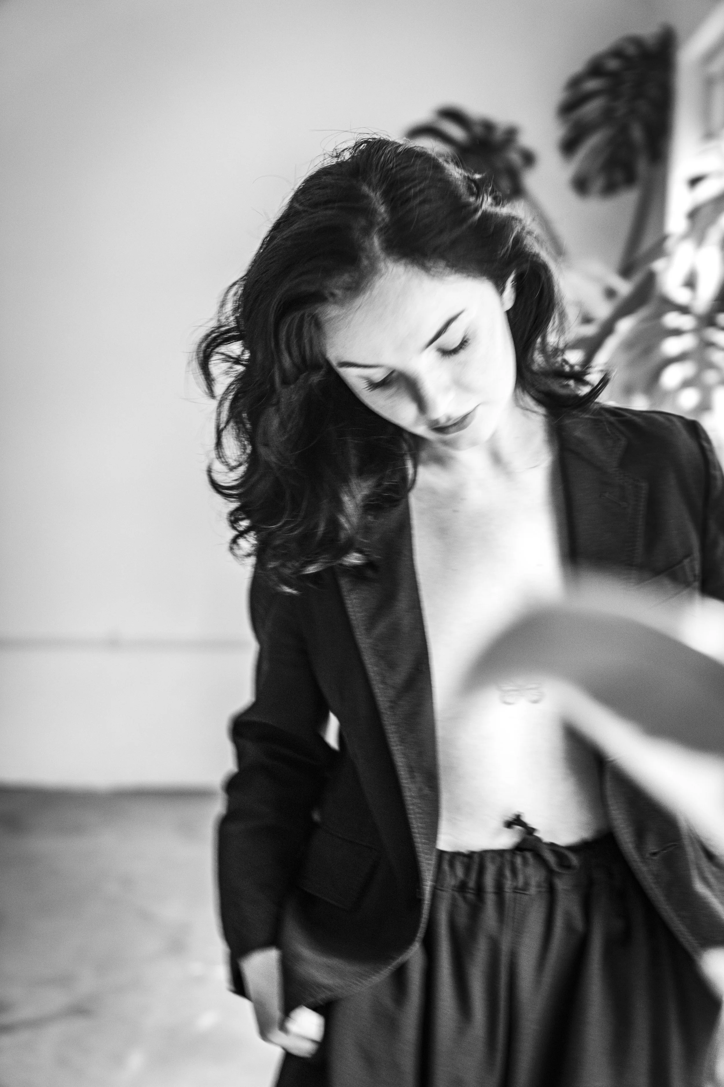 A woman with wavy dark hair looking down, dressed in a black jacket and striped pants, standing indoors with blurred plant in the background.