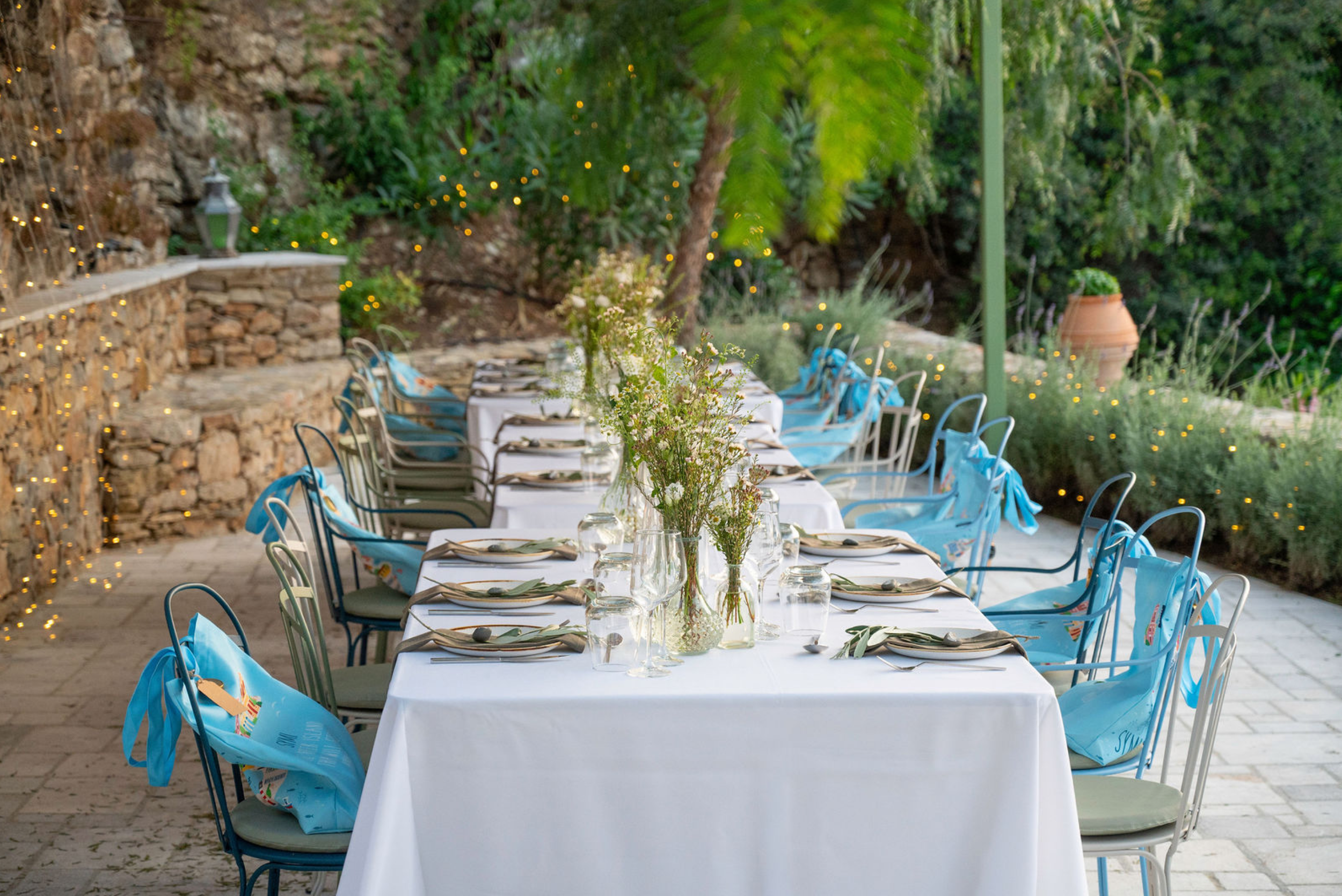 Outdoor dining setup with a long table covered in a white tablecloth, surrounded by chairs with blue sashes, set with plates, utensils, and glass jars of flowers, with a stone wall, greenery, and string lights in the background.