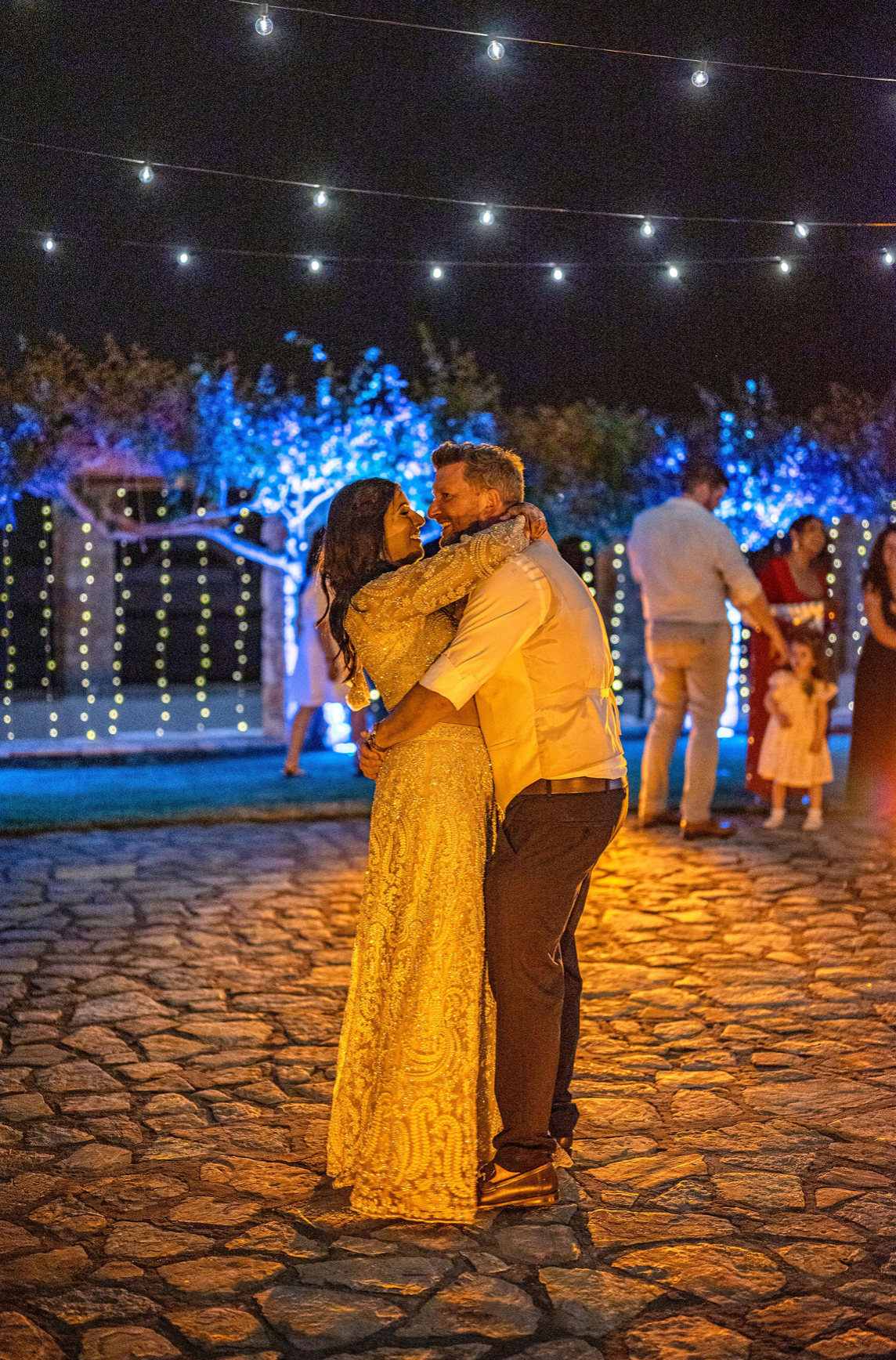 A couple dancing together outdoors at night, illuminated by string lights and decorative blue lights in the background, with other people visible in the distance.