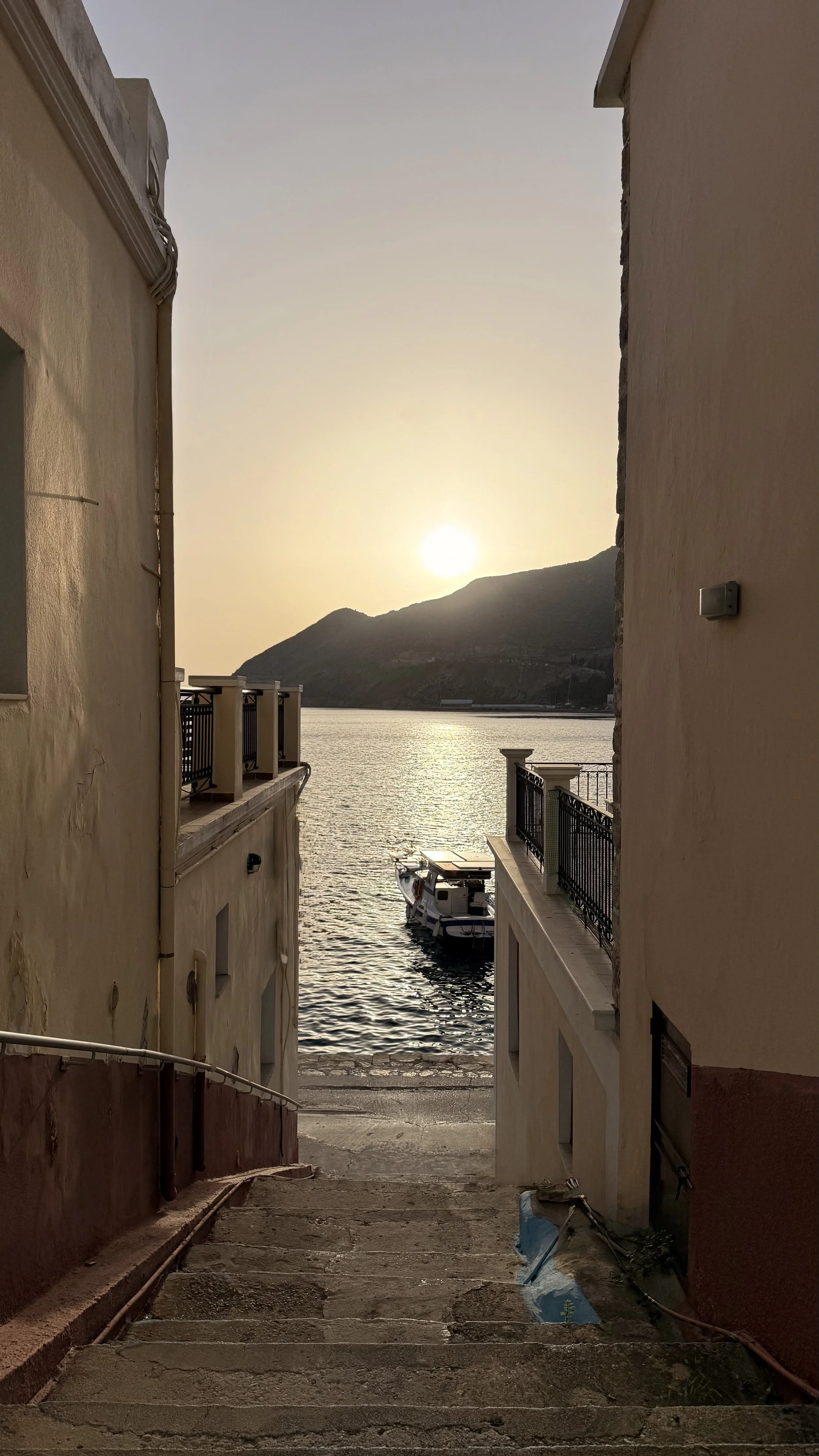 Narrow stairway leading down to a waterfront with a boat, framed by buildings on both sides, with a mountain and the setting sun in the background.