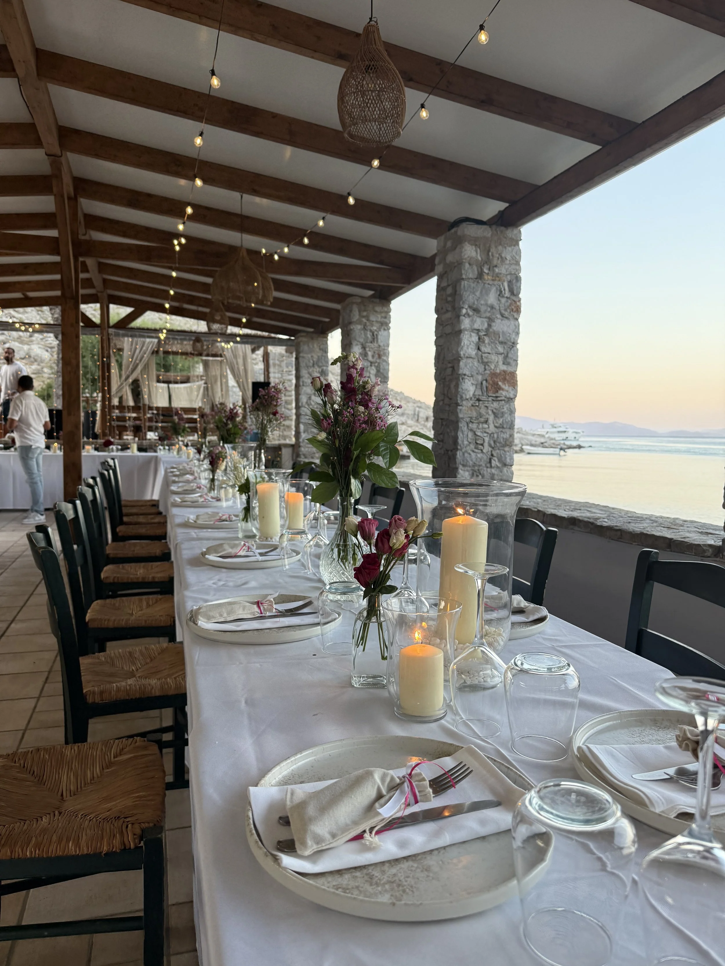 A banquet table set for an event with white tablecloth, floral centerpieces, candles, and candles in glass containers, overlooking a waterfront with boats at sunset. The setting is under a wooden roof with stone pillars, string lights, and wicker pen