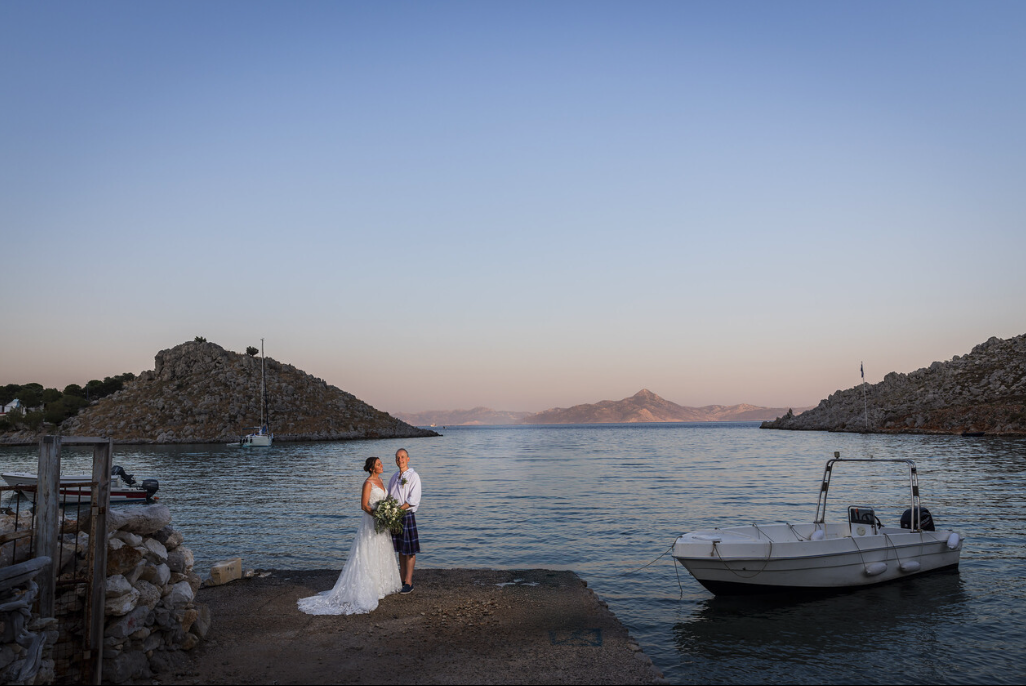 A bride and groom standing on a dock by the water, with boats and rocky hills in the background, during what appears to be a wedding or romantic occasion.