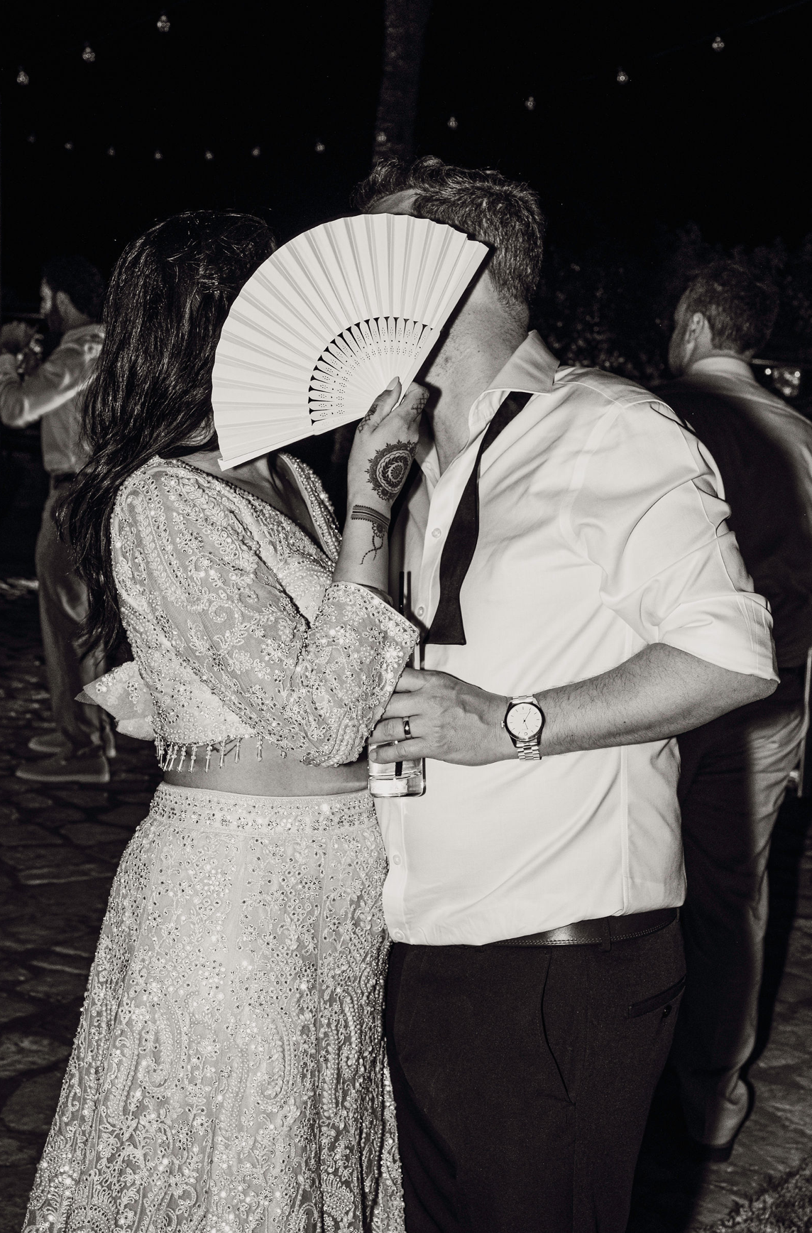 A couple dancing closely at a party, with the woman holding a fan and wearing an ornate dress, and the man wearing a white shirt with rolled sleeves and a watch, with other guests in the background.