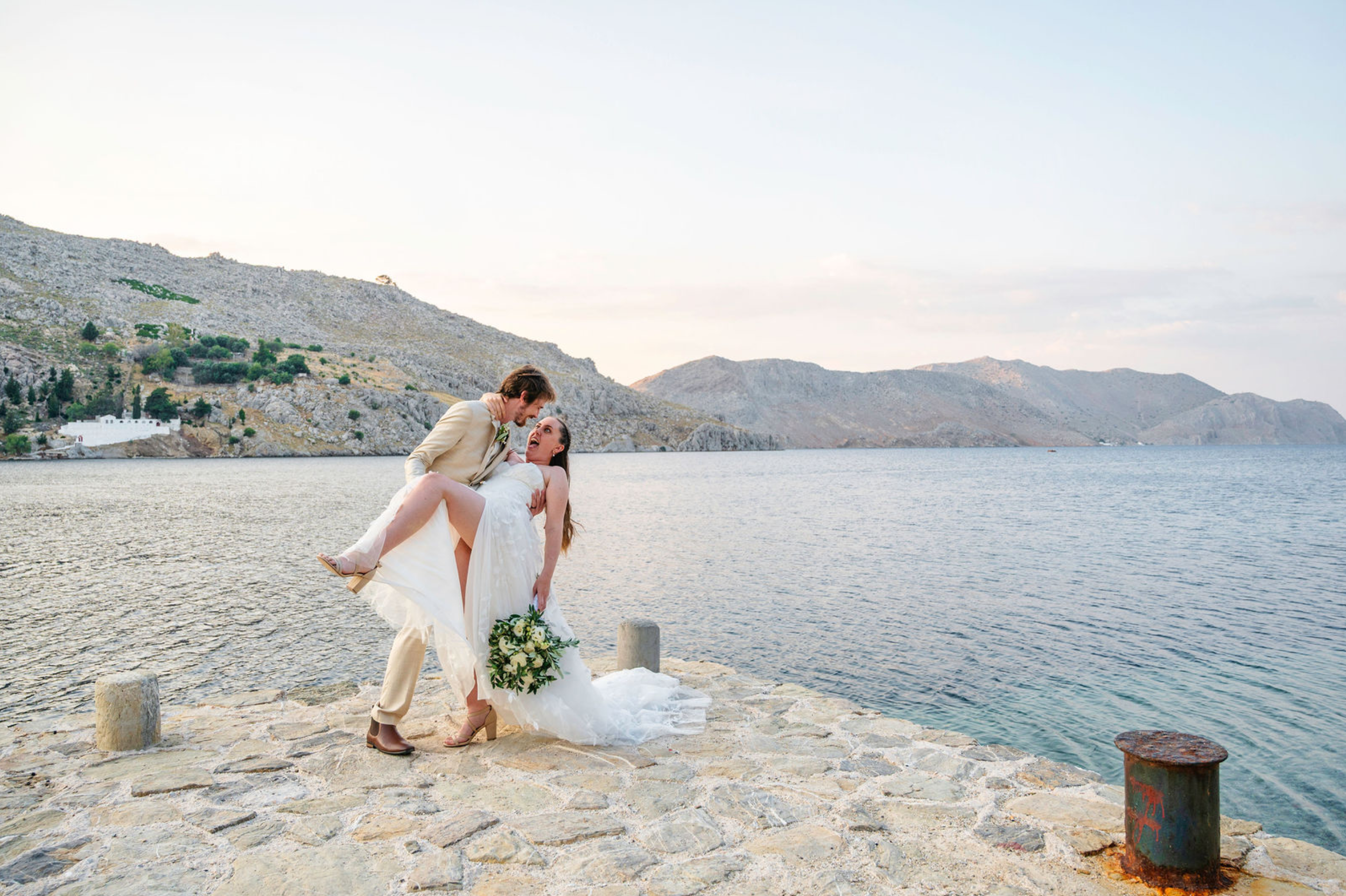 A bride and groom are by a body of water with mountains in the background. The groom is holding the bride, who is holding a bouquet of white flowers.