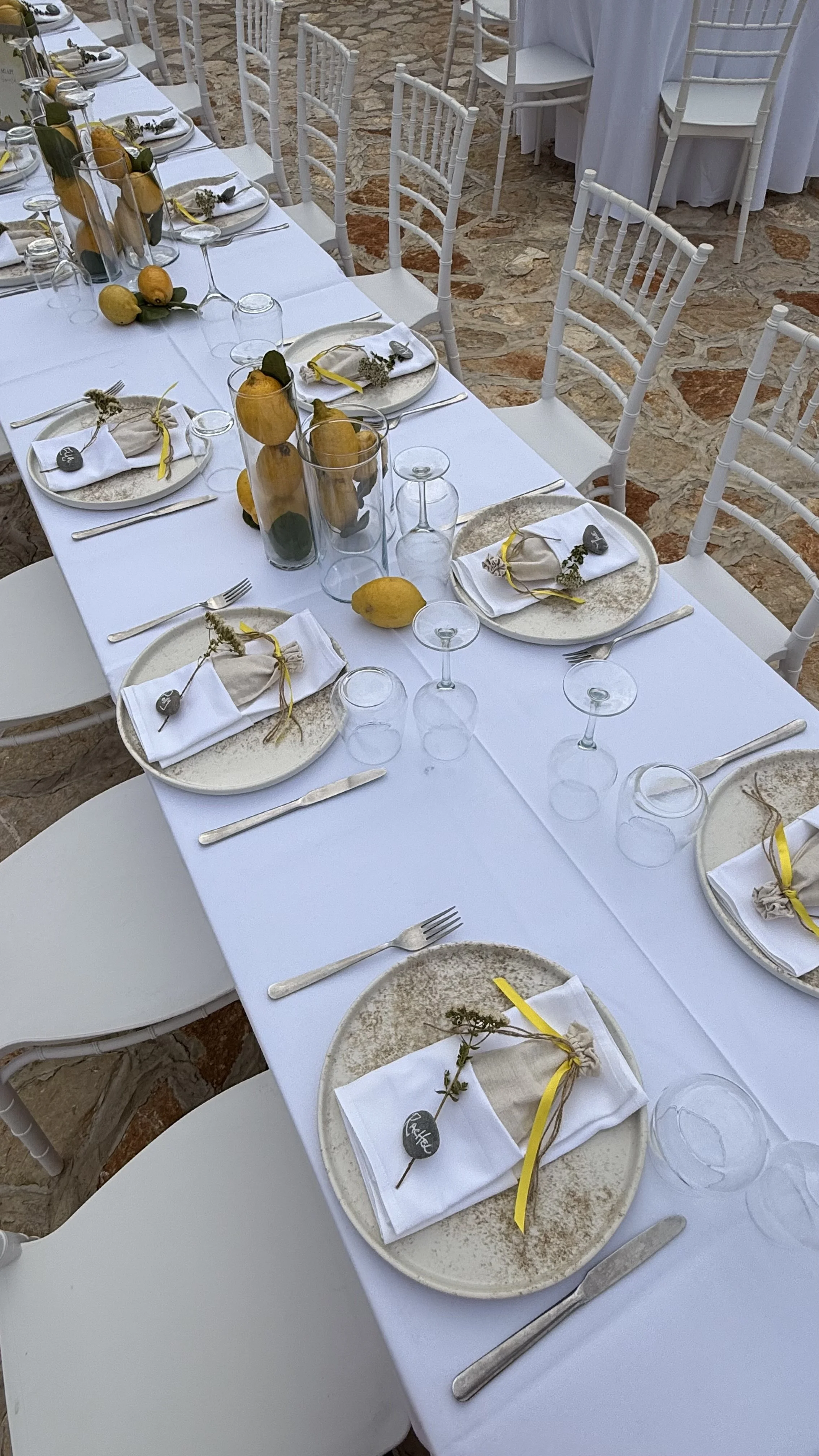 Elegant outdoor dining table set with white tablecloth, white chairs, decorative yellow butter pears, and lemon-colored ribbons tied around napkins with small chalkboard name tags.